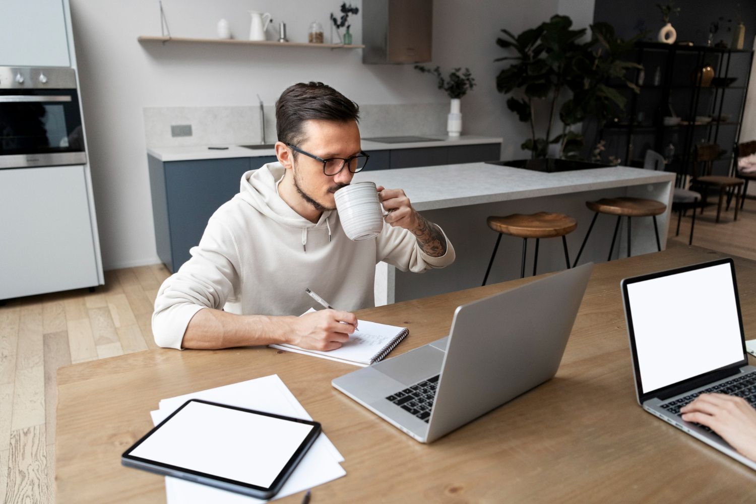 Man in glasses drinks from a mug while writing in a notebook at a table with a laptop and tablet.