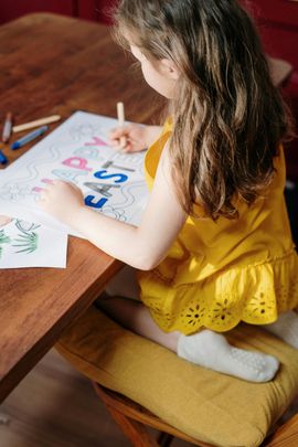 Child in a yellow dress drawing colorful letters at a wooden table.