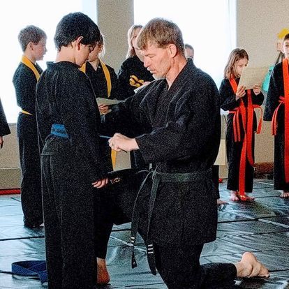 A man in a black karate uniform is kneeling down to help a young boy