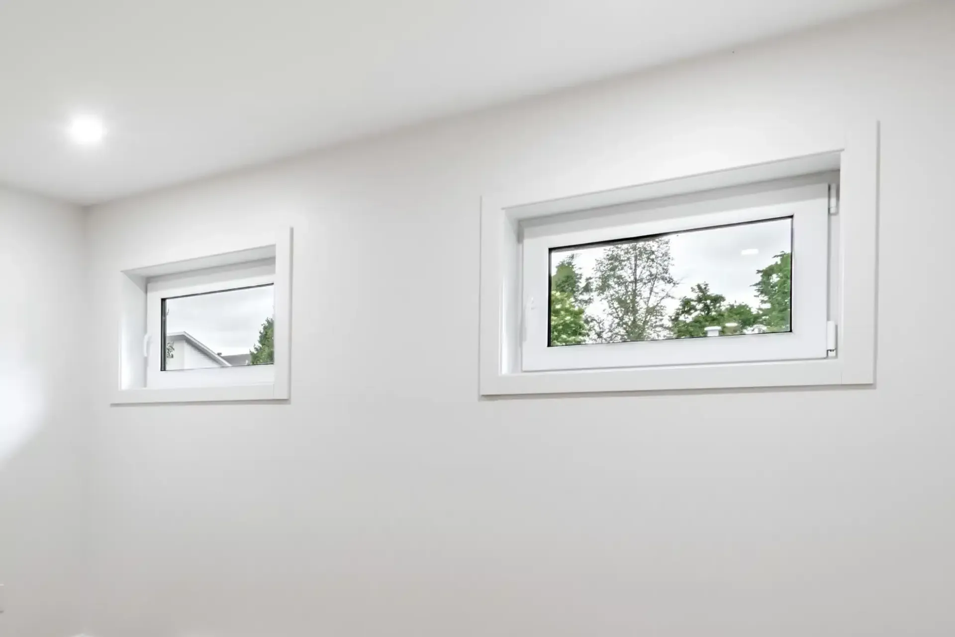 Two white-framed windows on a white wall; one larger than the other, overlooking trees and a roof.