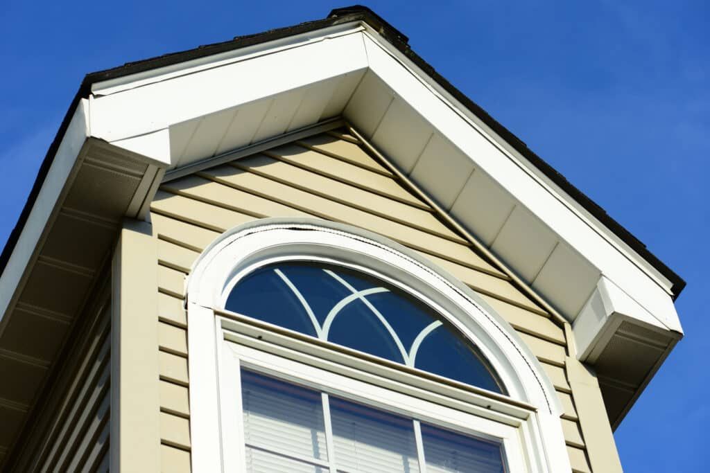Exterior view of a house with beige siding, white trim, and a semi-circular window under a clear blue sky.