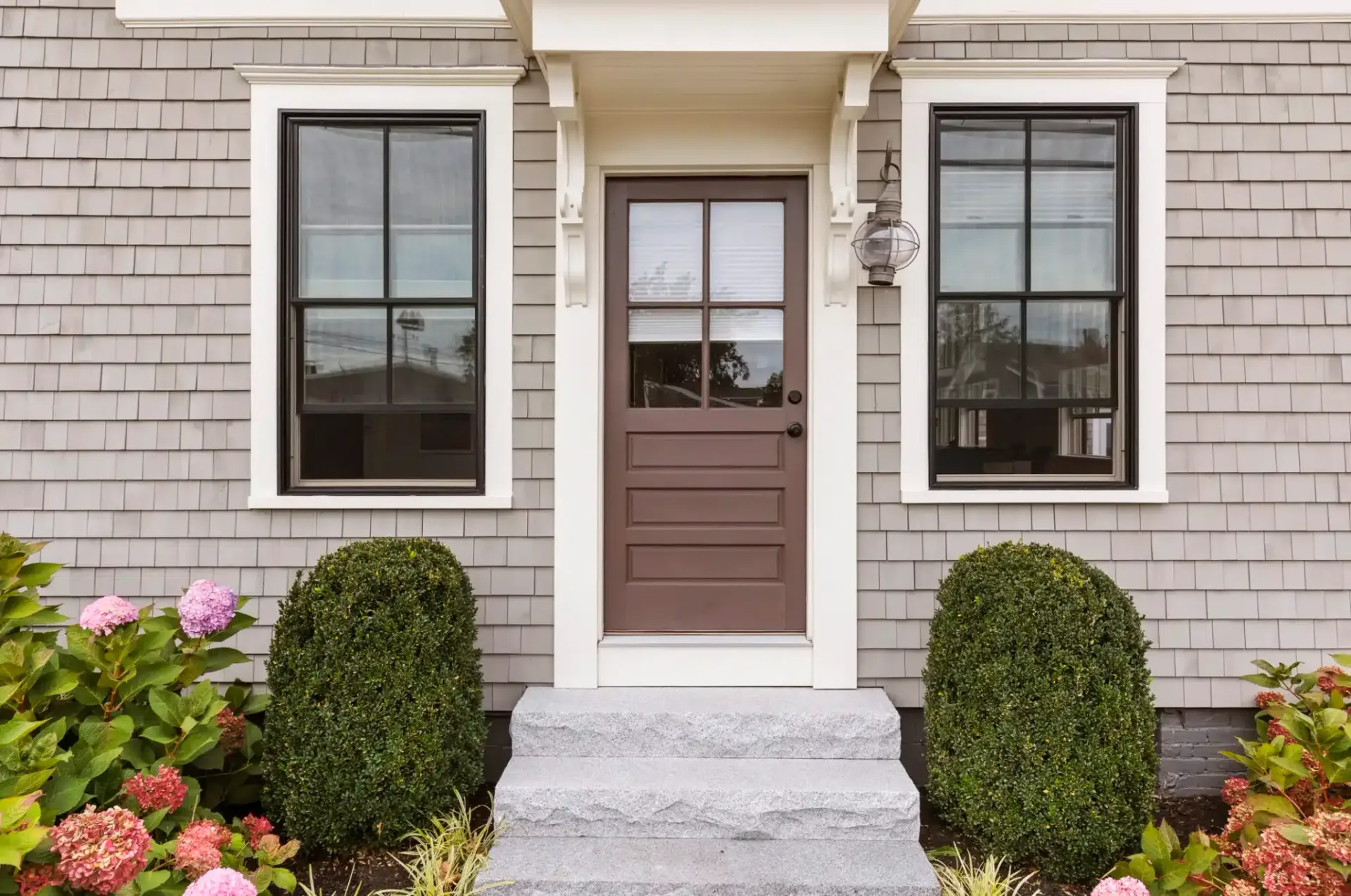 Brown door with windows flanked by black-framed windows, gray shingle siding, and stone steps, with bushes and flowers.