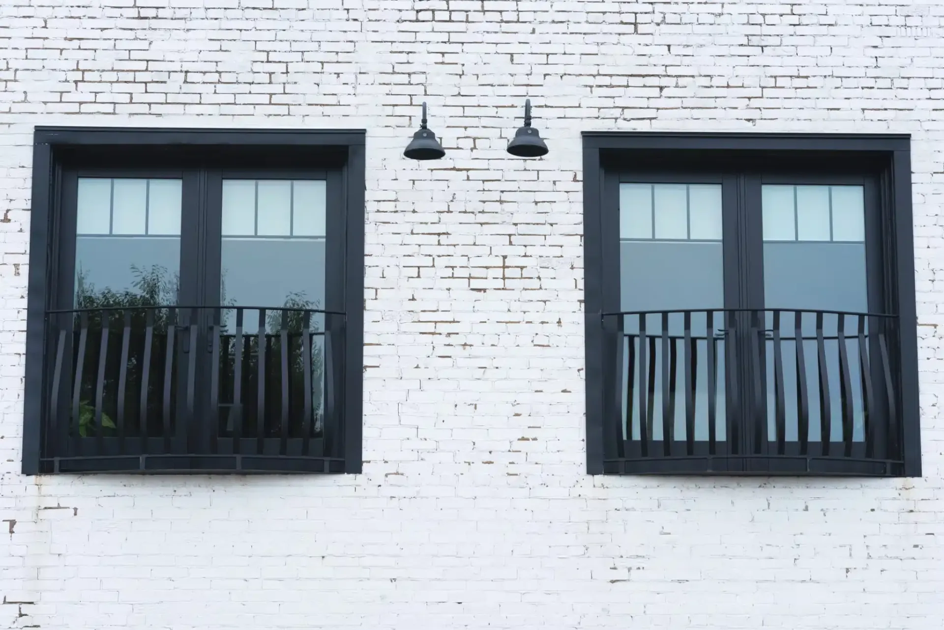 Two black-framed windows with small balconies on a white brick wall, two black lights above.