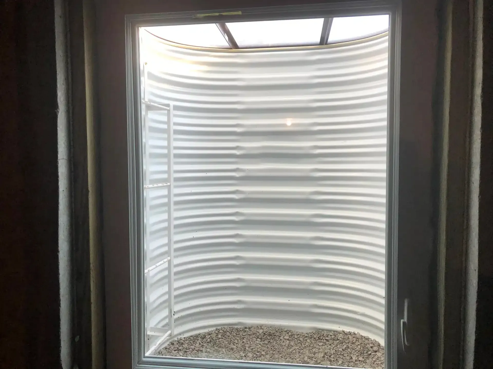 Basement window well with a ladder, covered by a translucent plastic dome, gravel at the bottom.