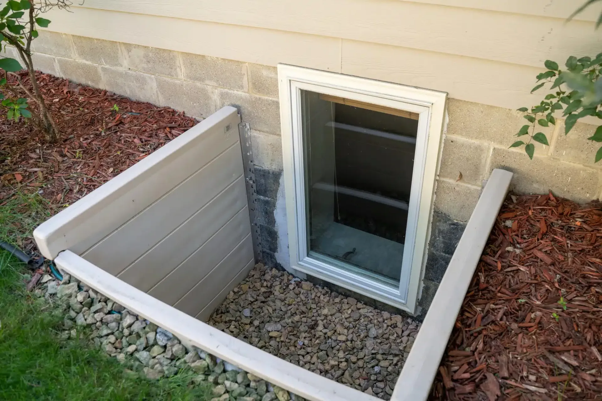 Egress Window well with white frame, surrounded by beige wood and gravel, in a brick foundation with red mulch.