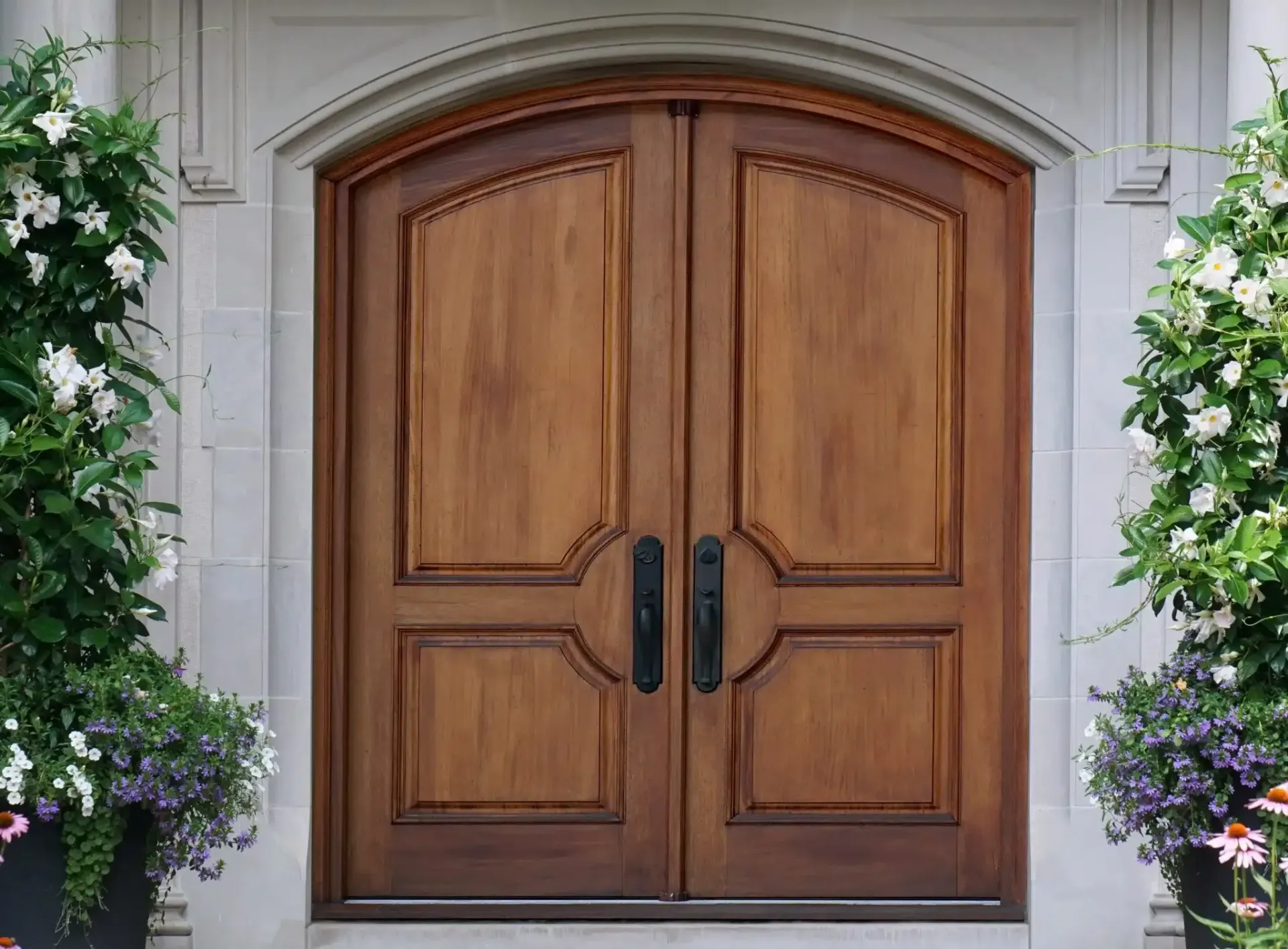 Wooden double doors with black handles, set in a stone entryway. Potted plants flank the entrance.