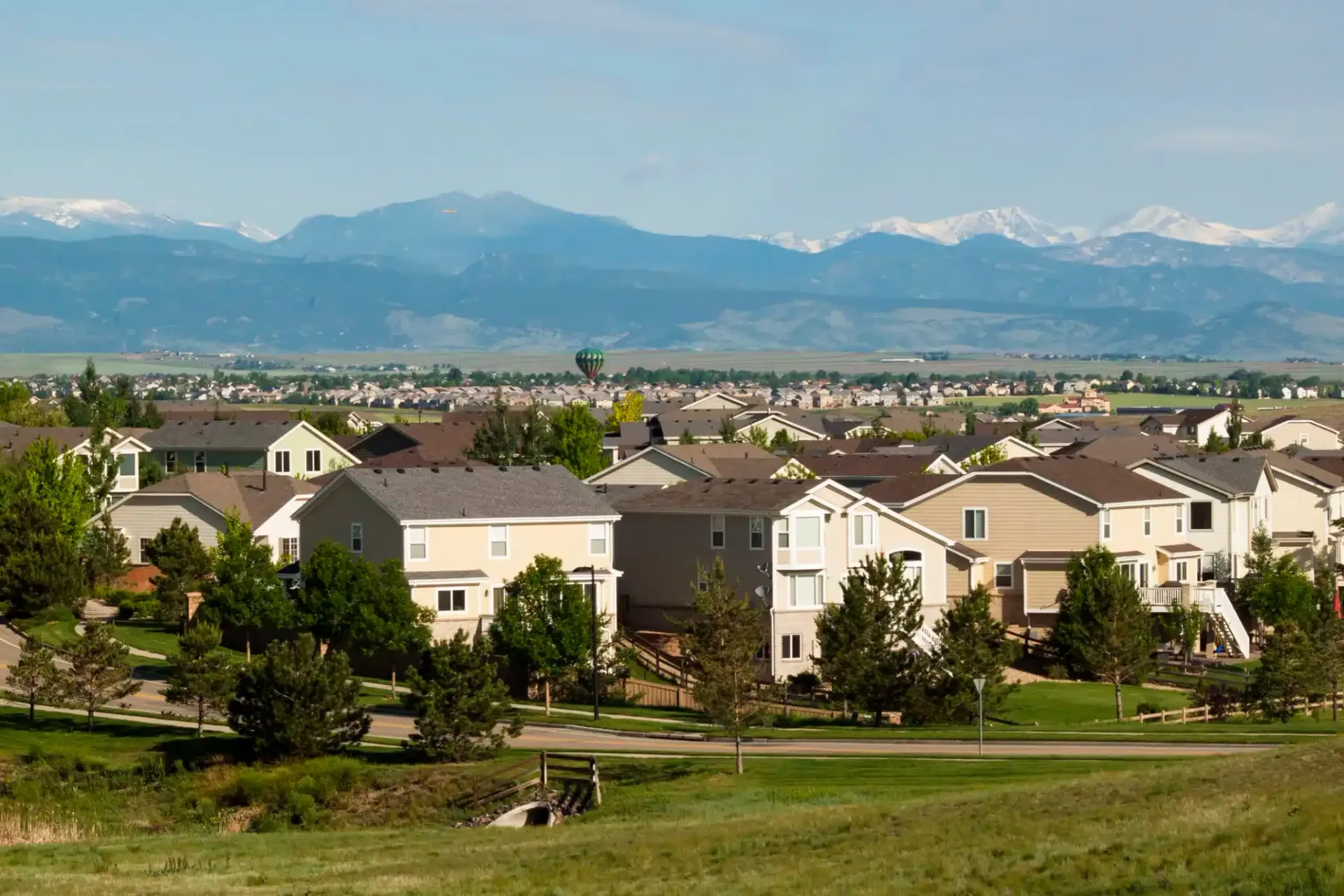 Suburban homes with green lawns and trees, mountains in the distance under a blue sky.