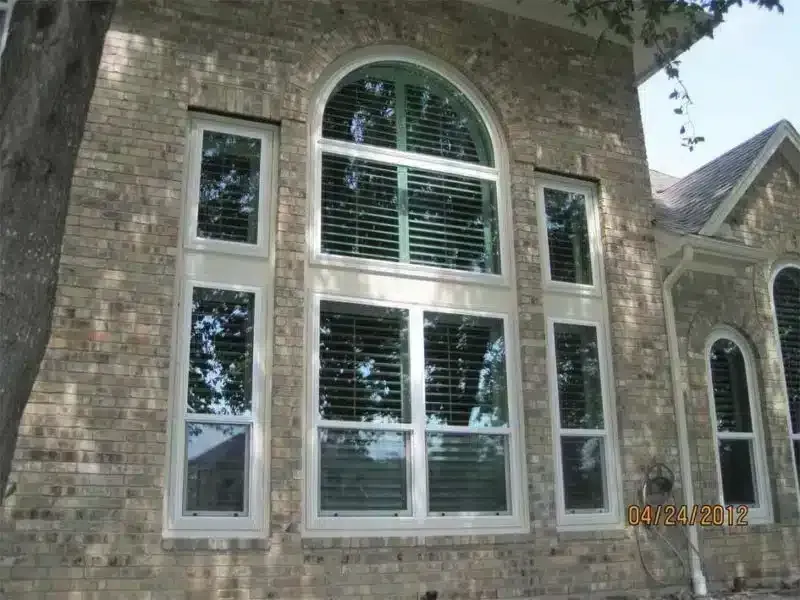 Exterior view of a brick house with large windows, including an arched one, with white frames and blinds.