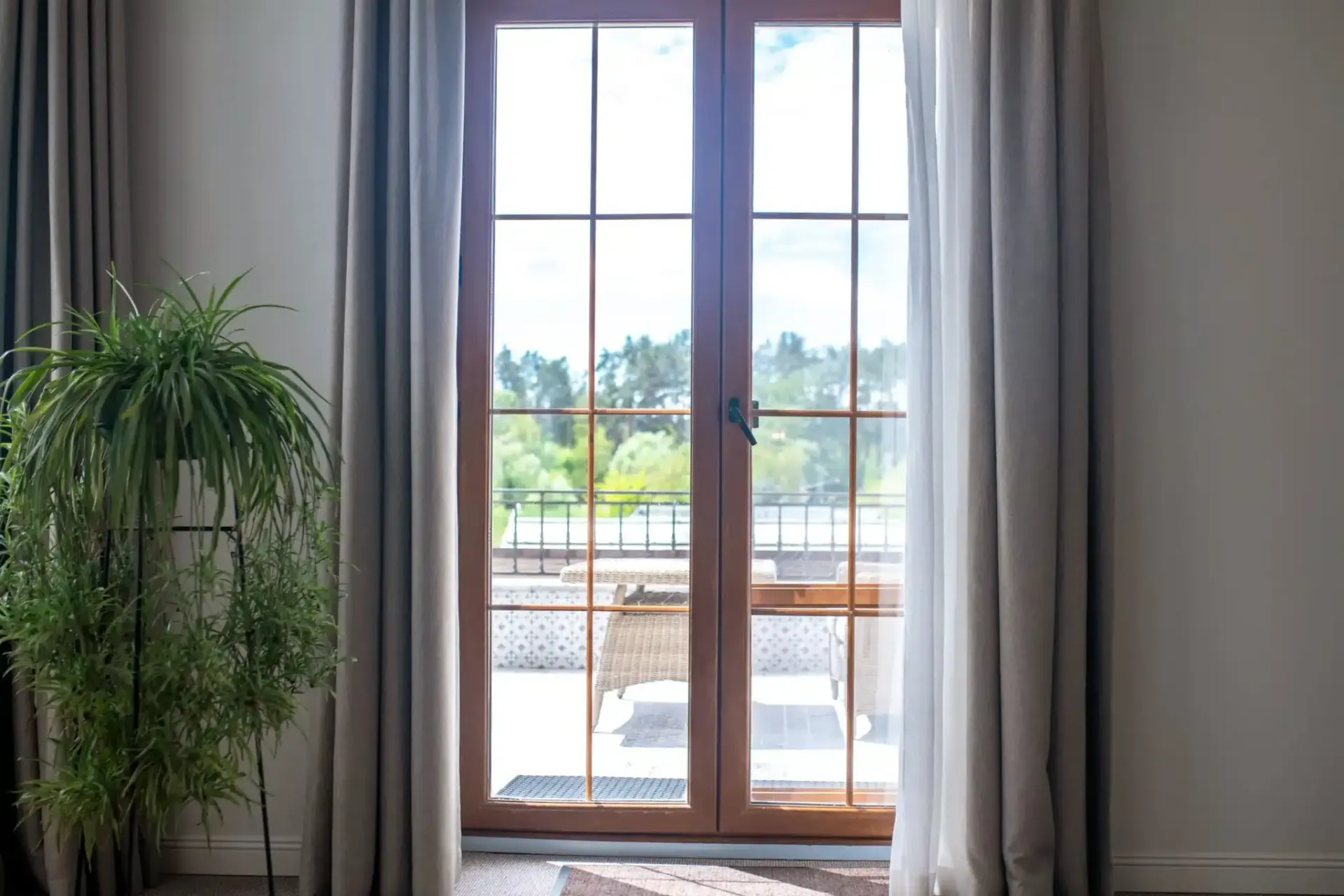 Wooden French doors with light-colored curtains and a leafy green plant. Exterior view with trees in the background.