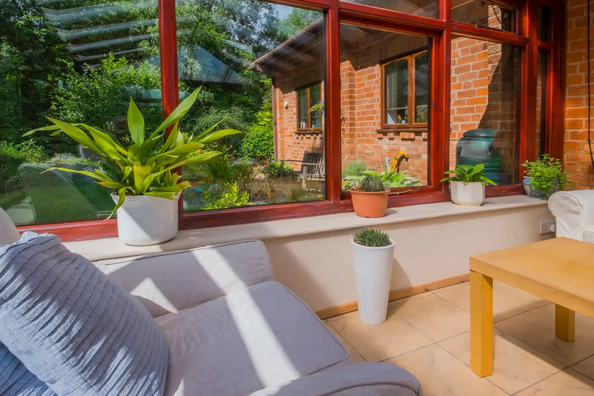Sunroom interior with plants on windowsill, couch, and wooden table. View to brick house and greenery outside.