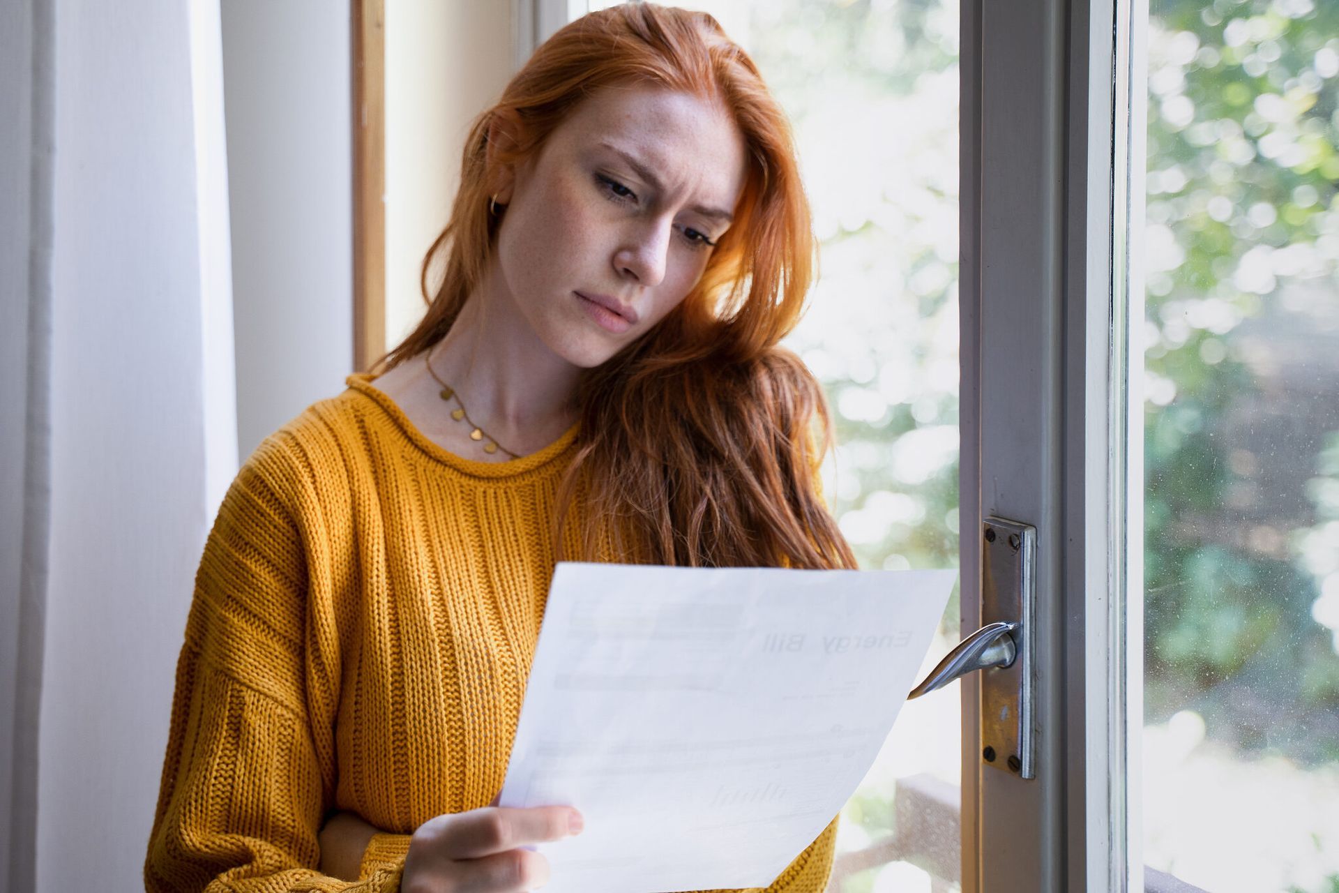 Woman with red hair looks concerned, holding a letter, near a window.