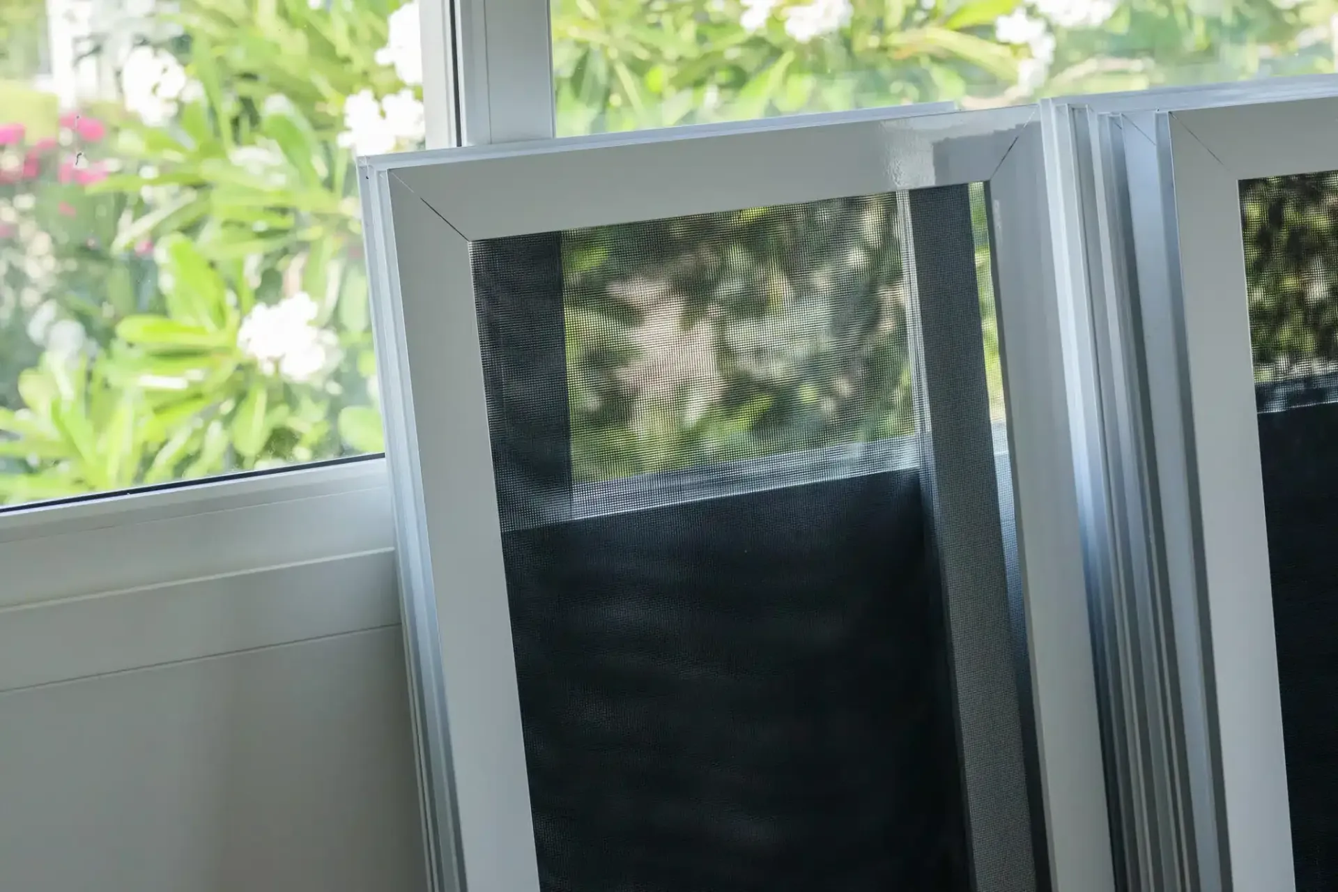 White-framed window screens stacked near a window, with a blurry outdoor view through the window and screens.