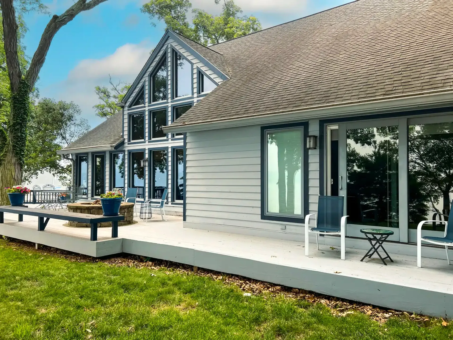 House with large windows and a deck overlooking water. Blue and gray color scheme, sunny day.