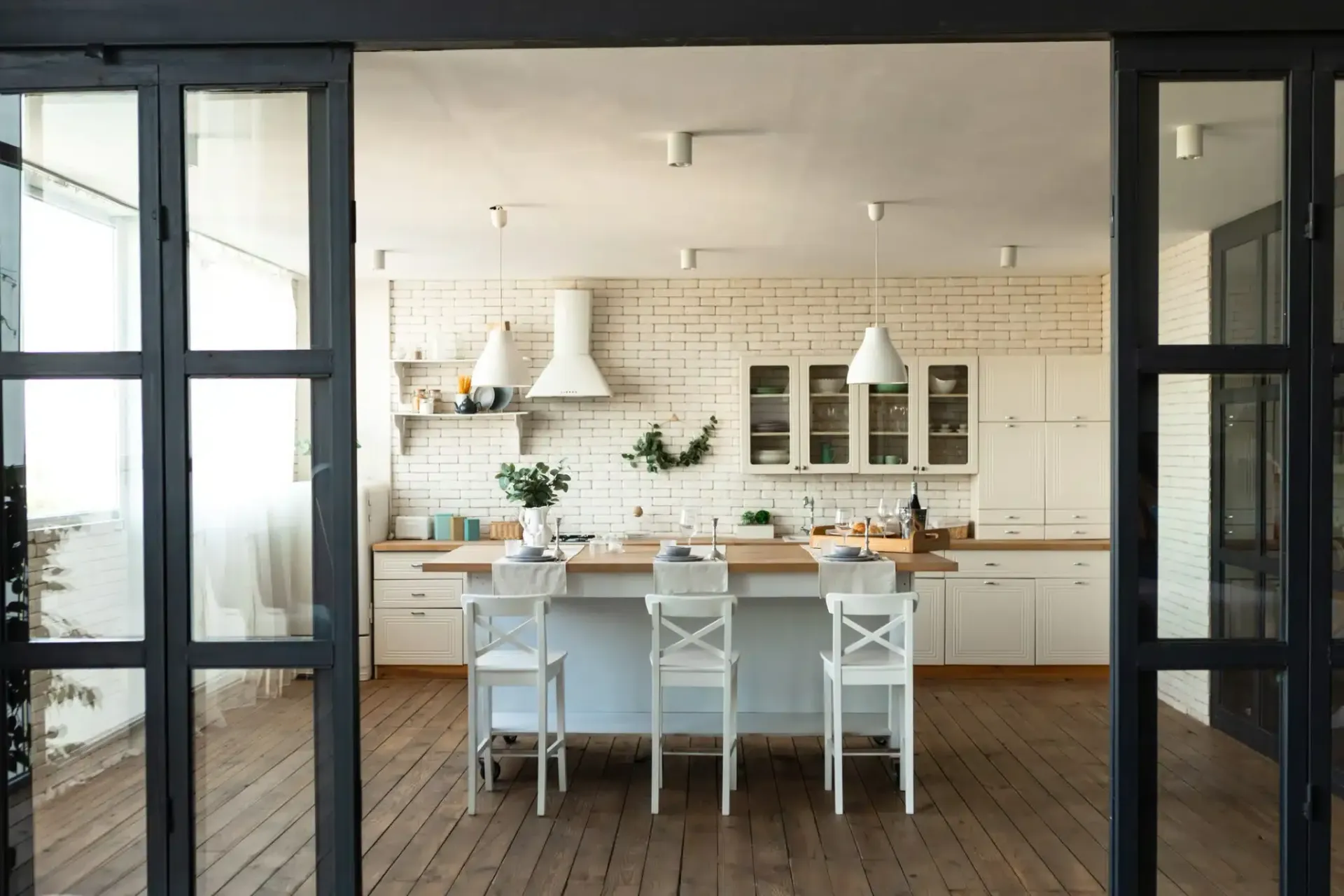 Bright kitchen with white cabinets, island with stools, and brick wall. Black framed doors.