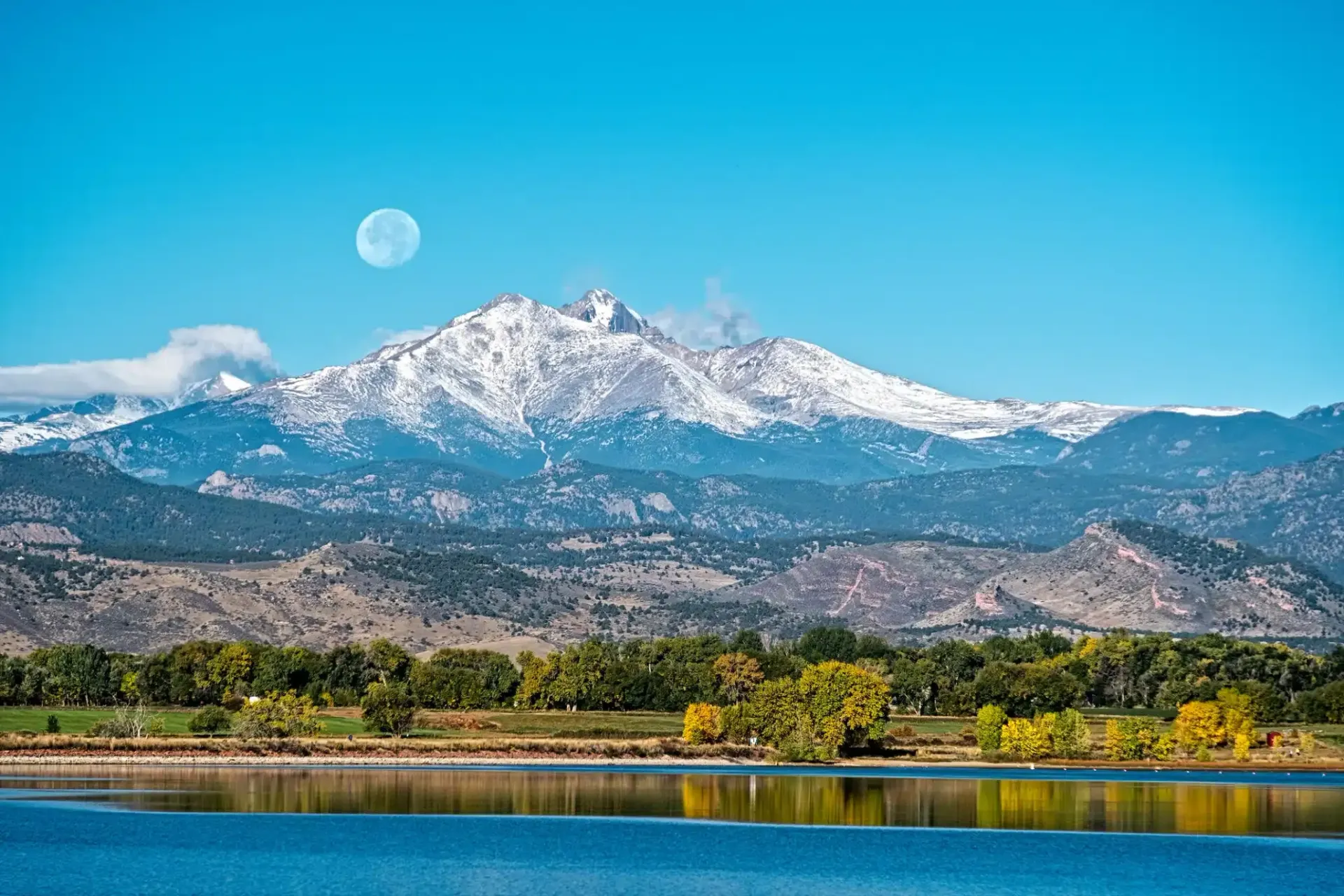 Snow-capped mountains with a lake in the foreground under a bright blue sky with a moon.