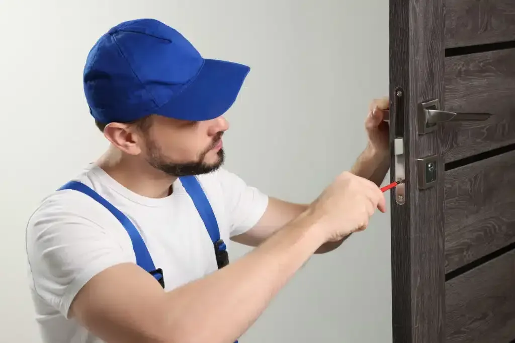 Locksmith in blue hat and overalls, fixing door lock with screwdriver.