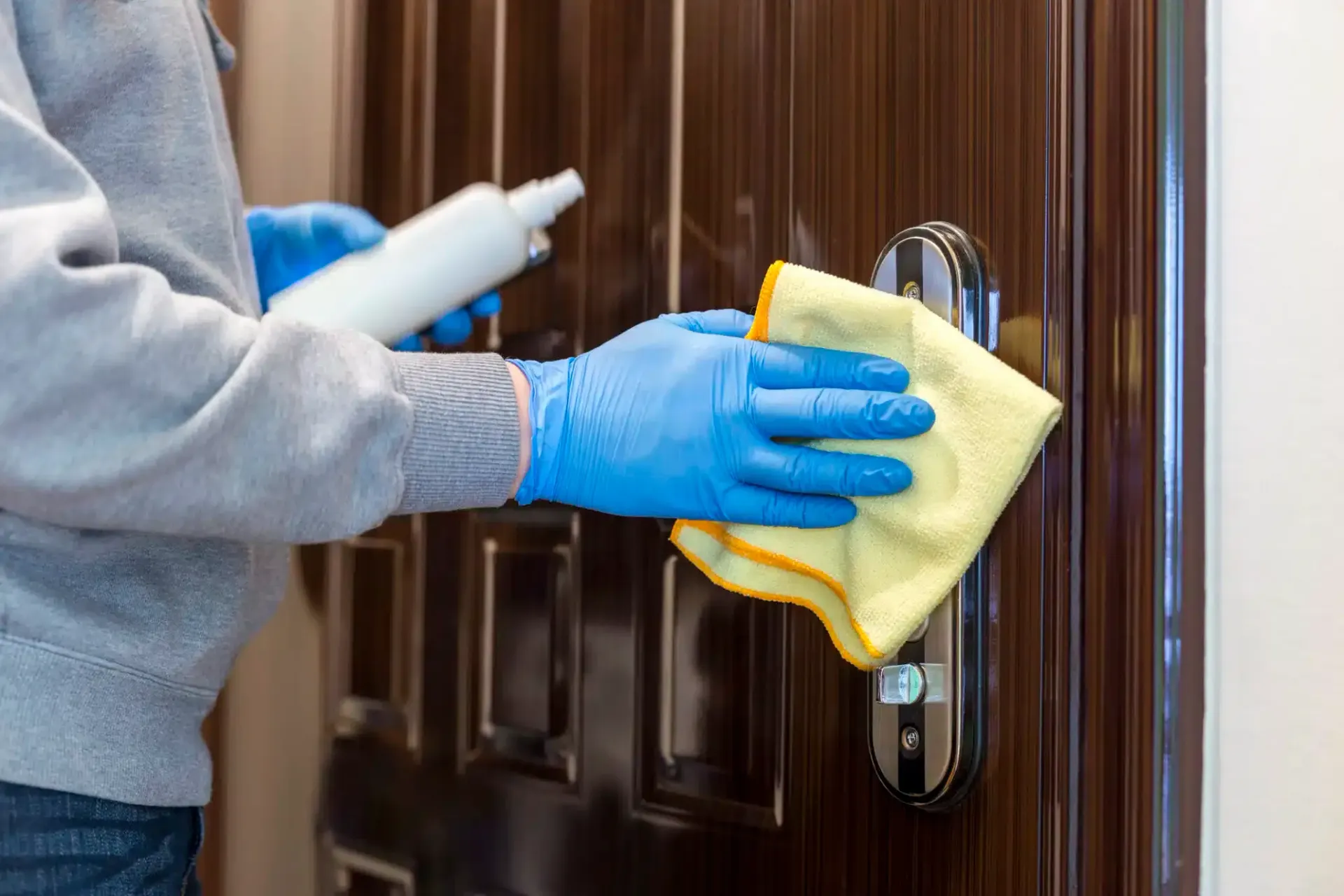 Person in blue gloves sanitizing a dark brown door handle with a spray bottle and yellow cloth.
