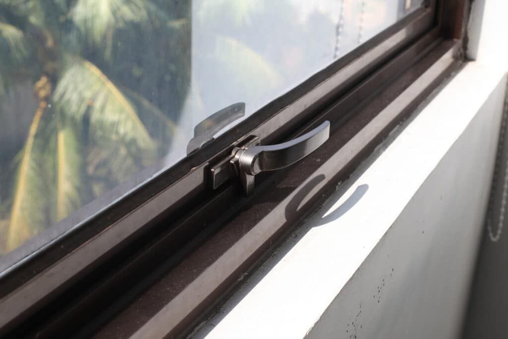 Close-up of a window latch on a brown frame, with a bright window view of green palm trees in the background.