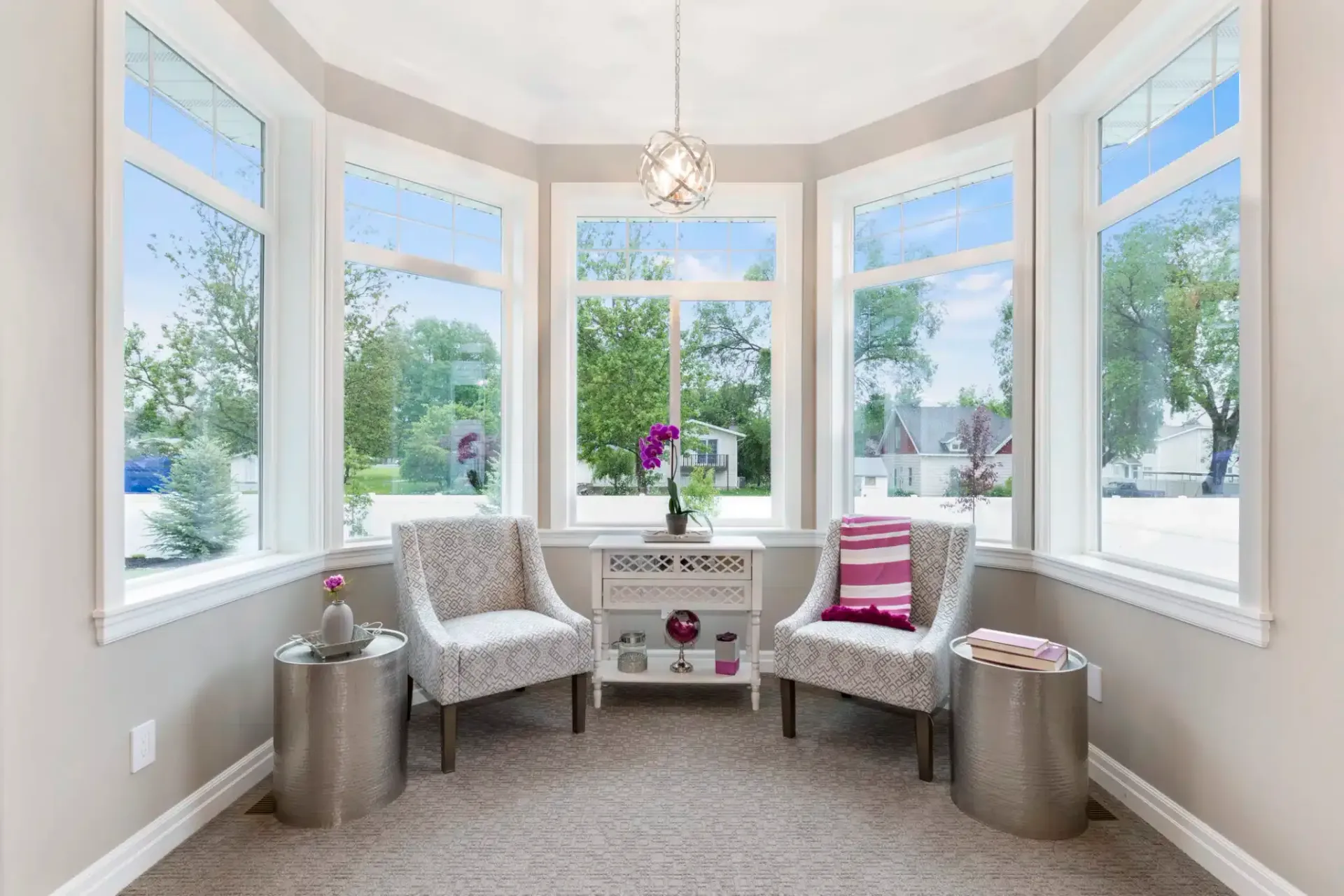 Sunroom with bay windows, two patterned chairs, small table with decor, silver stools, and an area rug.