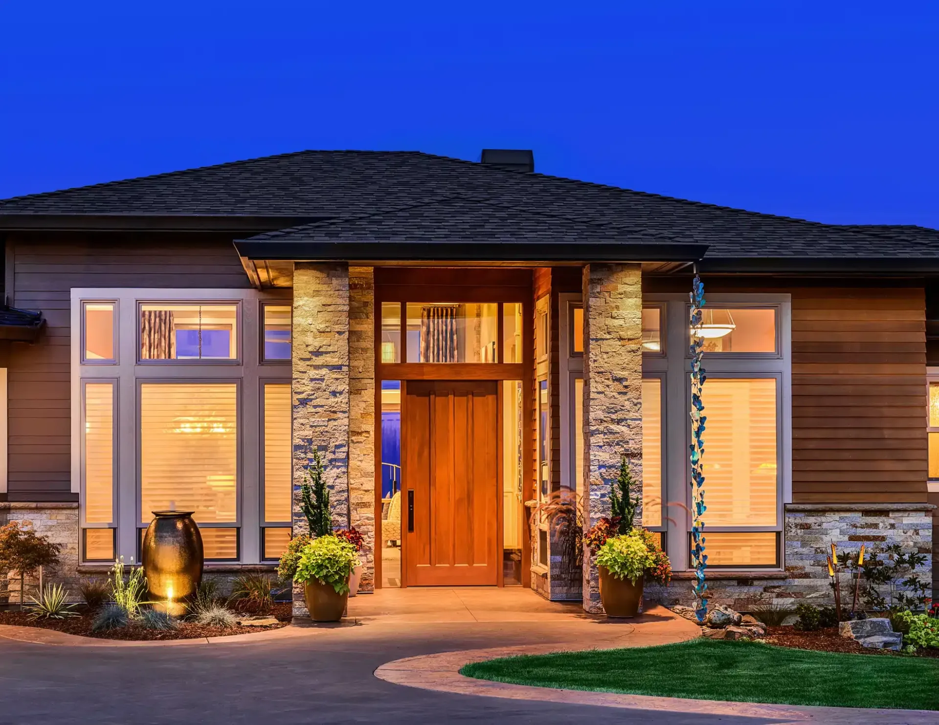 Modern house exterior with wooden door, stone pillars, and large windows at dusk.