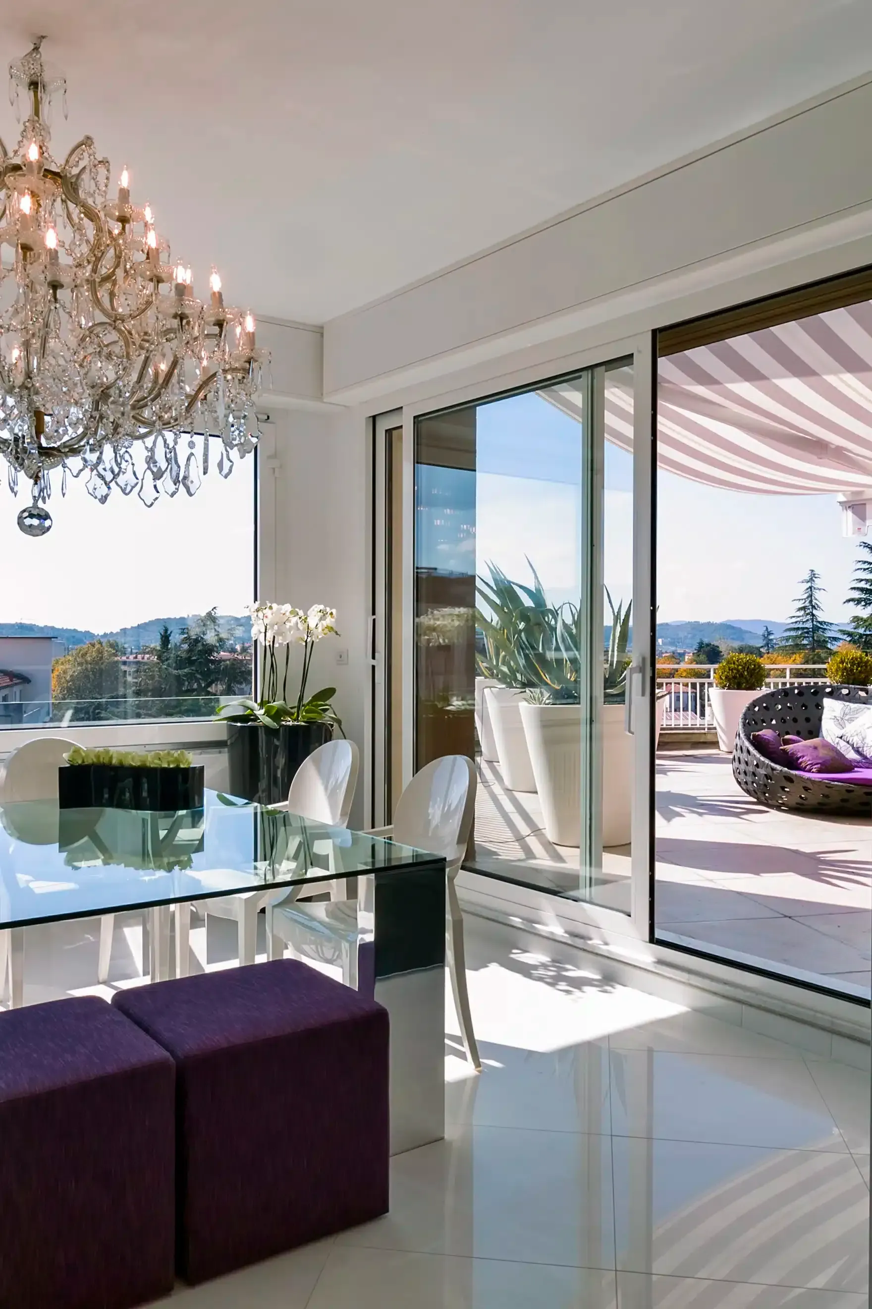 Dining room with glass table, purple ottomans, crystal chandelier, and open sliding door to a balcony.