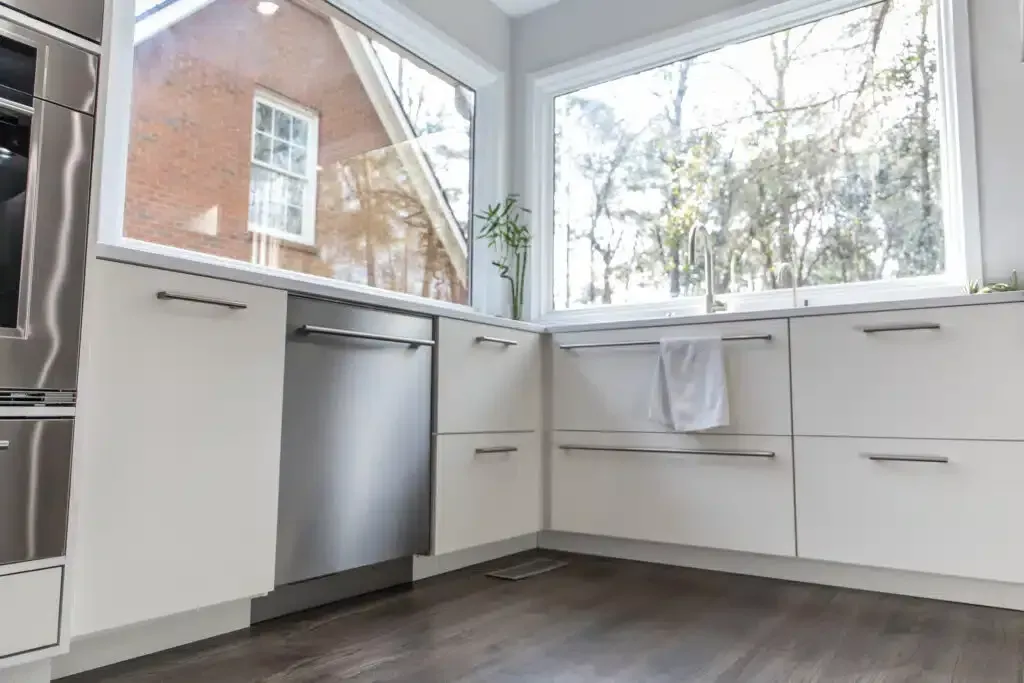 Stainless steel dishwasher and off-white cabinets in a kitchen with large windows and a view of trees.
