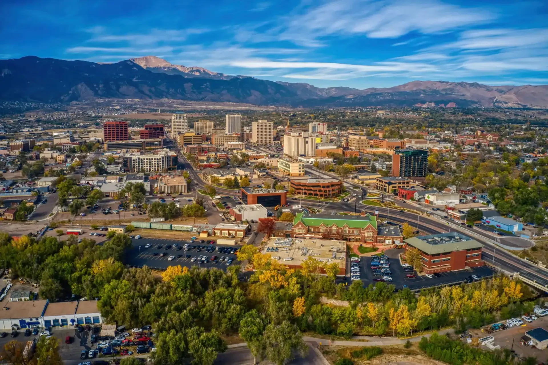 Cityscape with buildings and mountains in the background under a blue sky. Autumn trees in foreground.