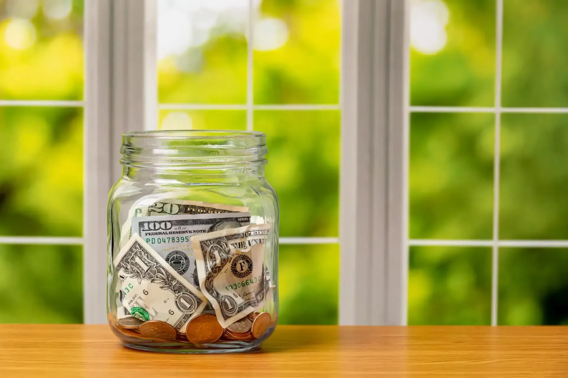 Glass jar filled with money on a table, with a window and green foliage in the background.