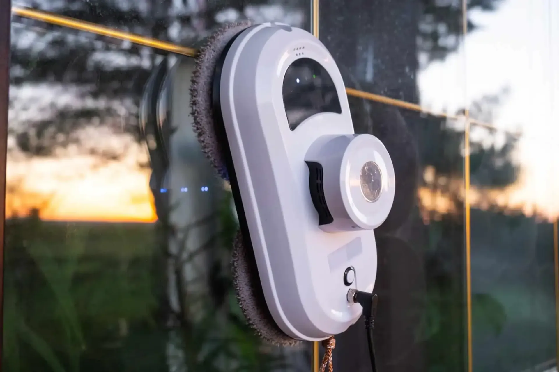 Window-cleaning robot on a glass pane, gray and white with circular cleaning pads. Sunset and foliage visible outside.