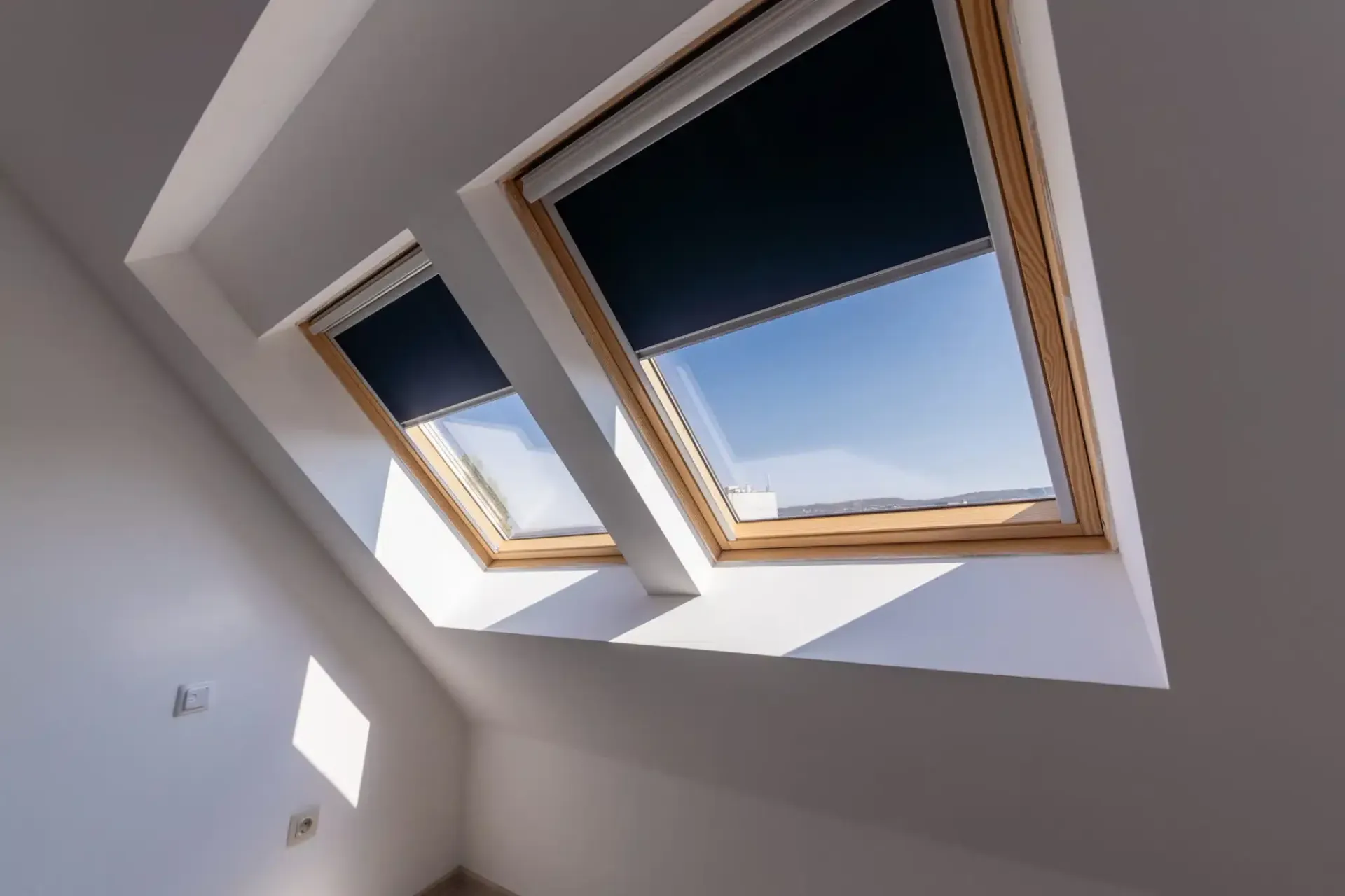 Two angled skylights with dark shades in a white-walled room, bright blue sky visible.