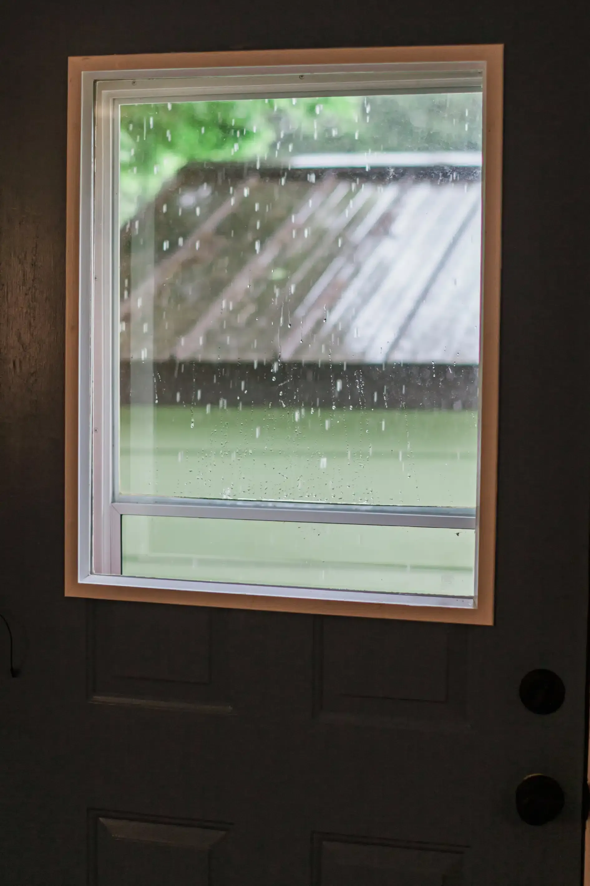 Gray door with rectangular window. Raindrops on the window obscure a view of a deck and green foliage.
