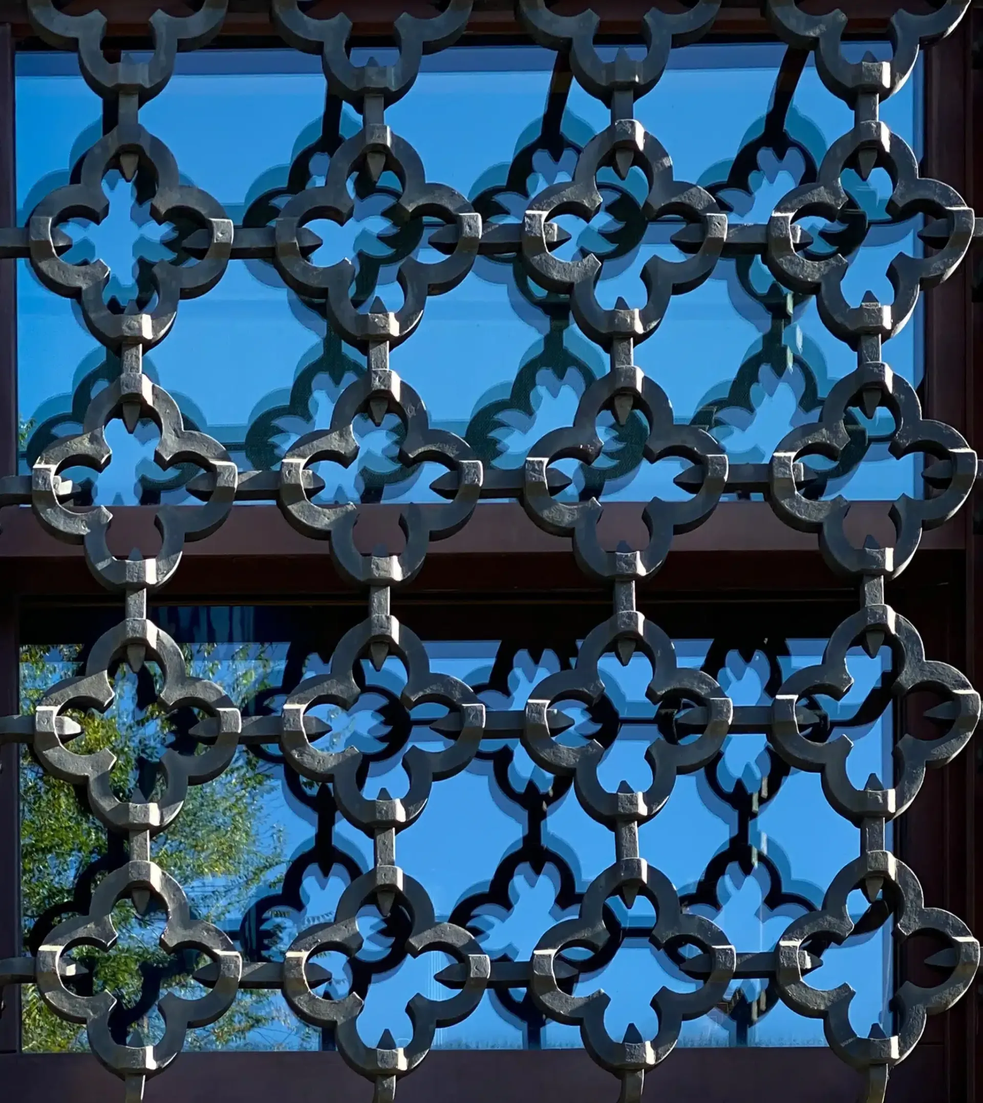 Black decorative metal window grille against a blue sky, reflecting trees.
