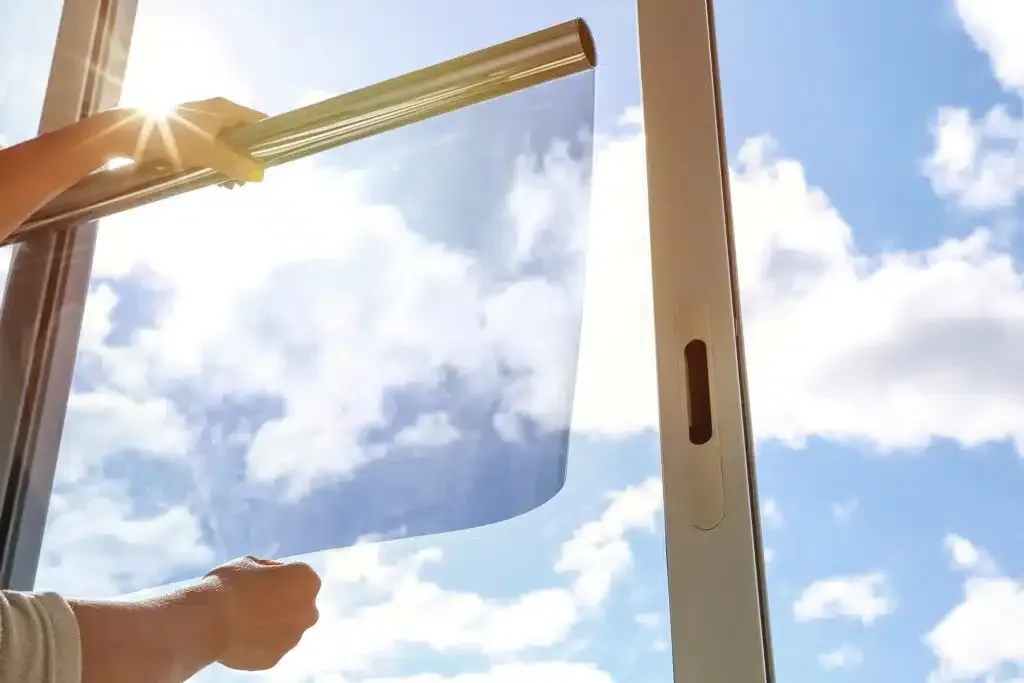 Person applying window film to a window, with blue sky visible through the film and window.