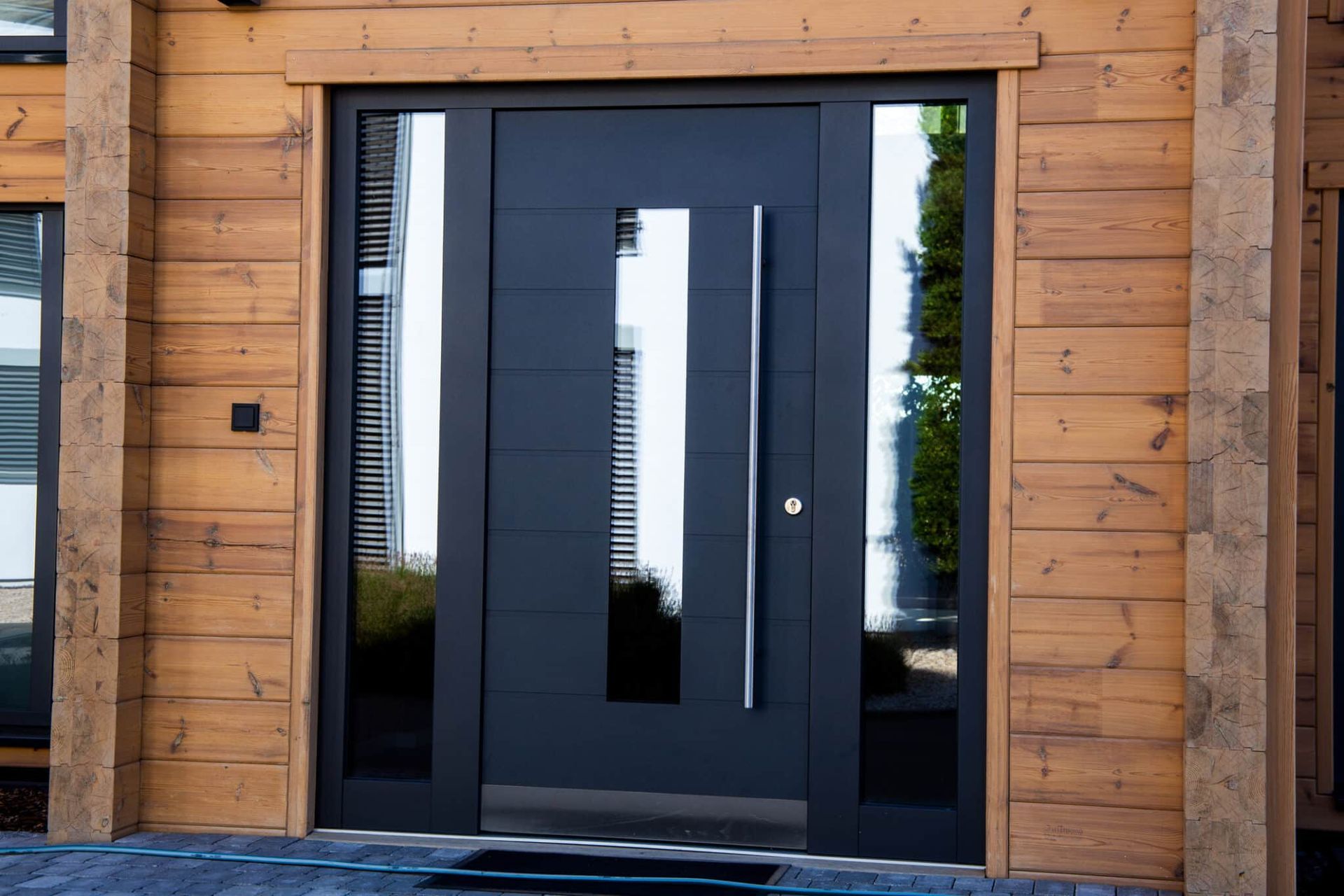Black front door with long vertical handle, glass panel, set into wooden siding.