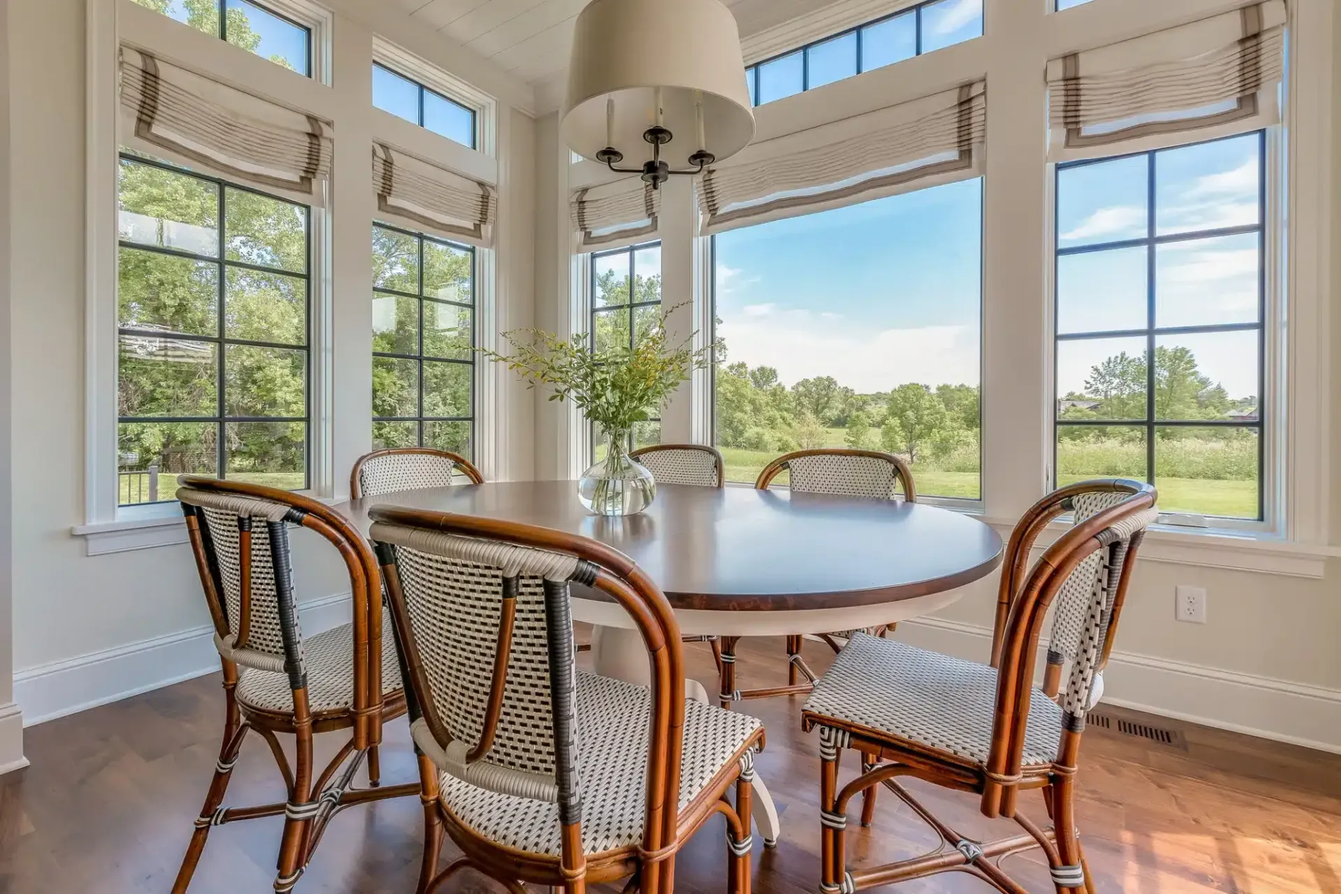 Round dining table with six wicker chairs in a sunroom with large windows overlooking a field.