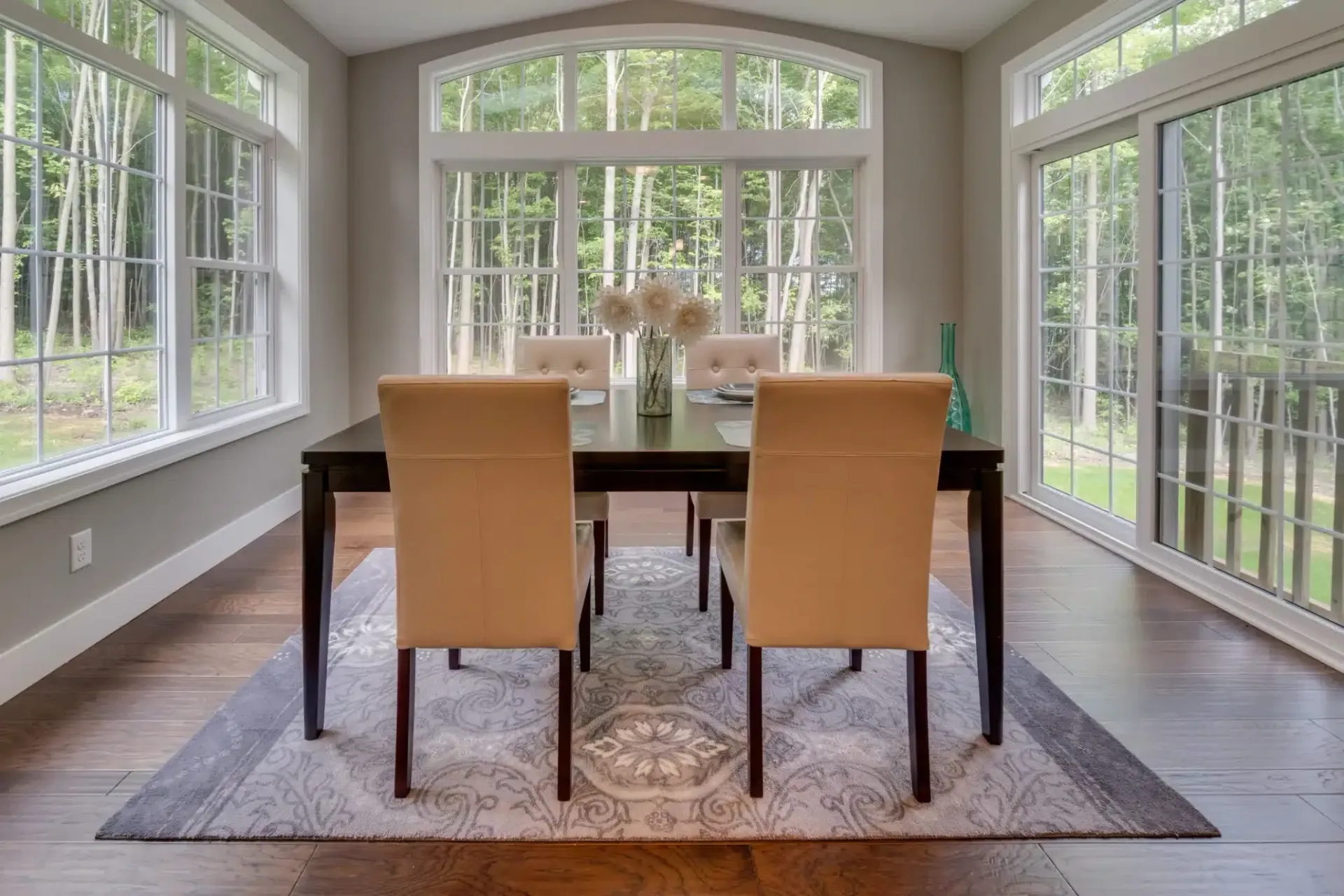 Dining room with dark table, beige chairs, and large windows overlooking trees.