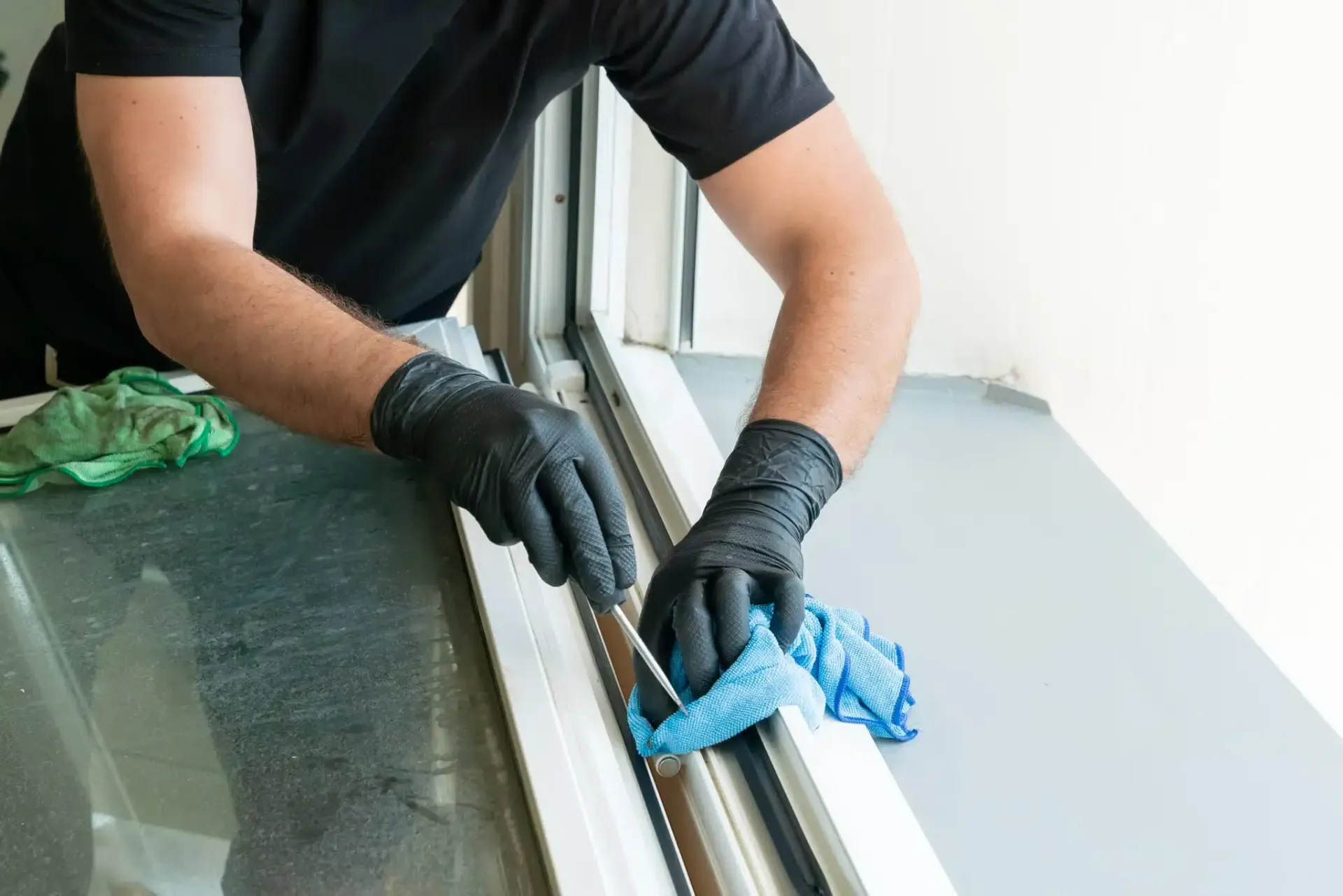 Person wearing black gloves cleaning a window track with a blue cloth.