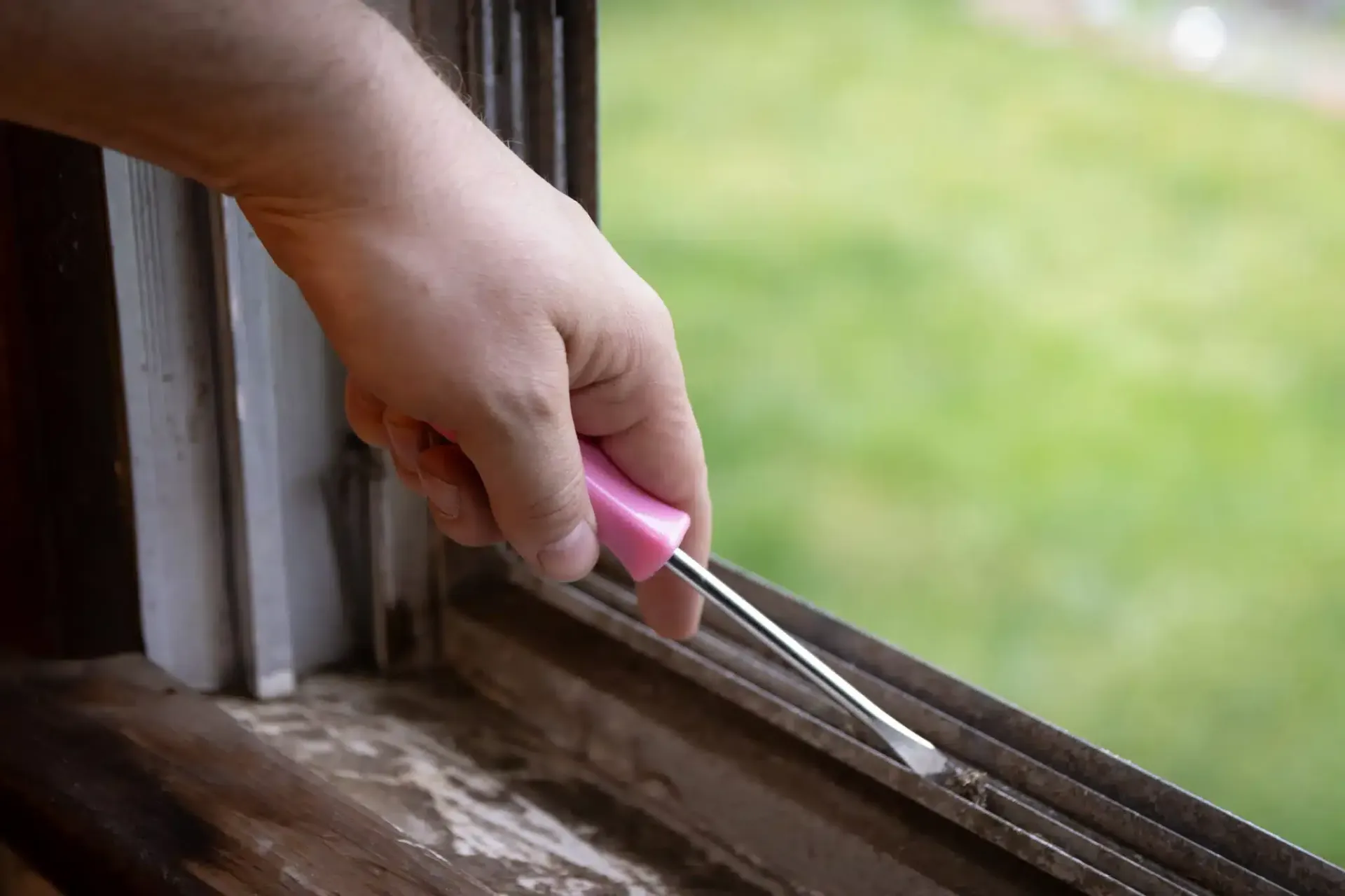 Hand using a pink tool to clean a dirty window frame, with a blurry green background.