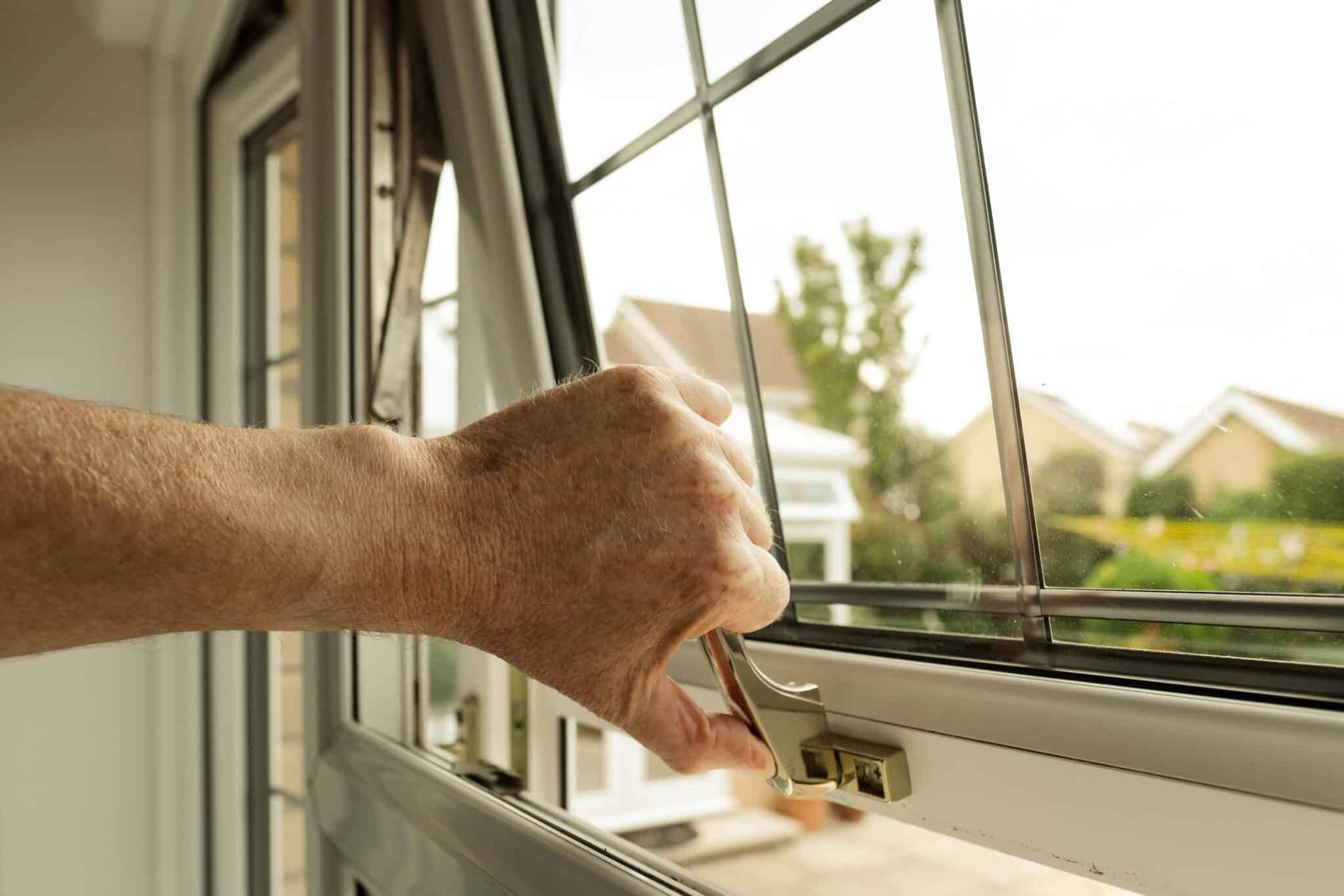 Hand opening a casement window with a lever, showing a suburban street in the background.