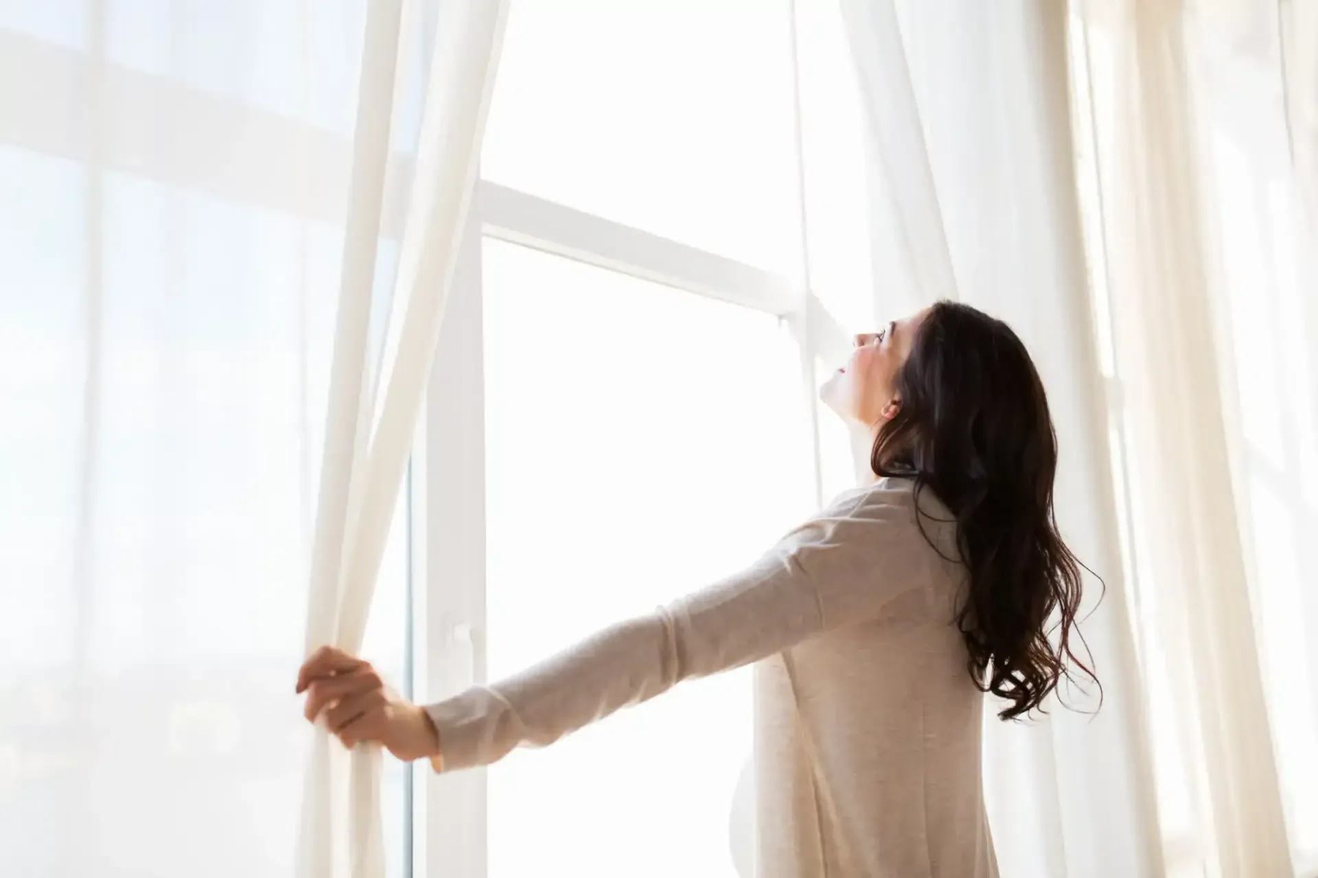 Woman opening curtains, looking up with a hopeful expression at a bright window.