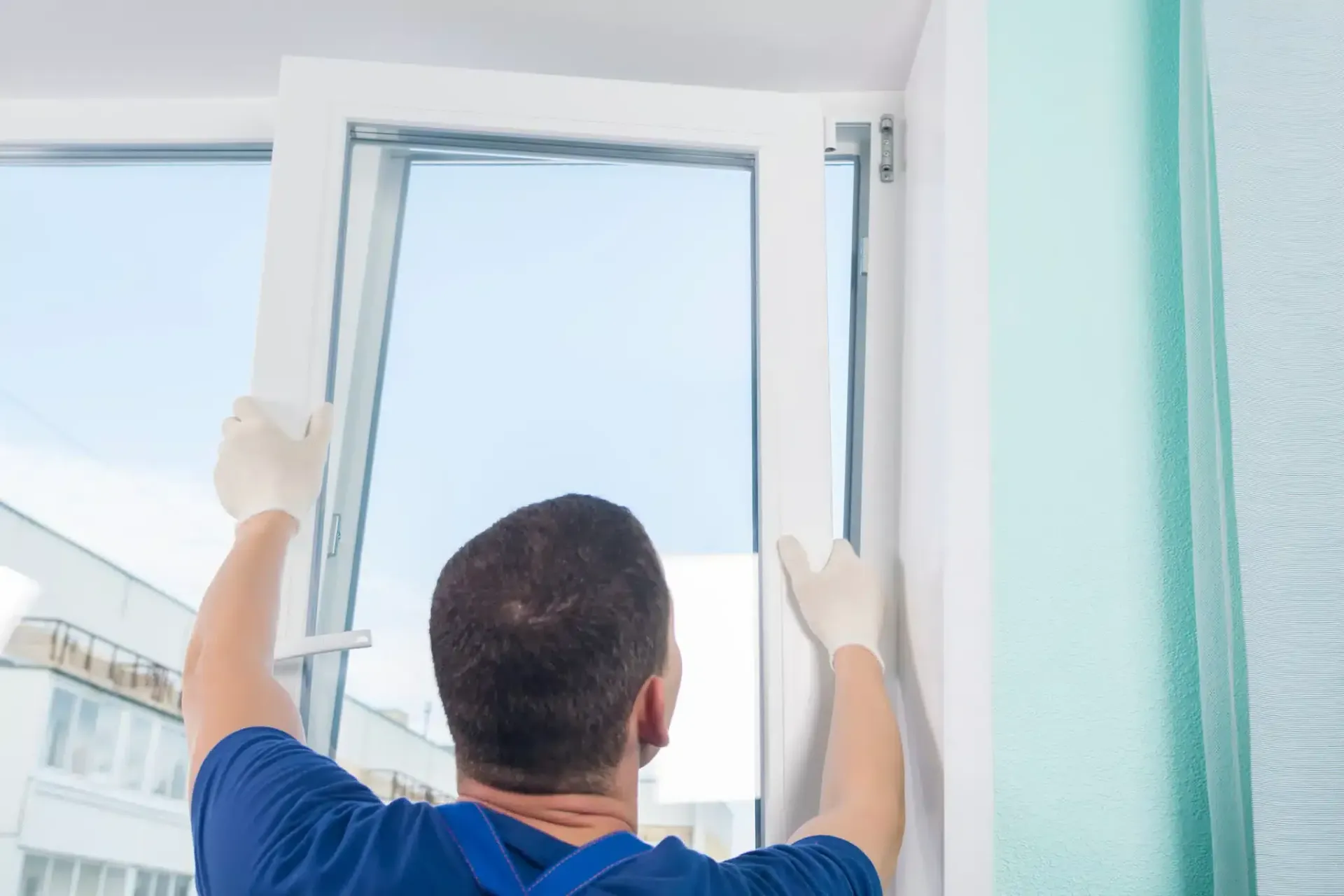 Person installing a white window, wearing gloves and blue overalls, in a light green room.