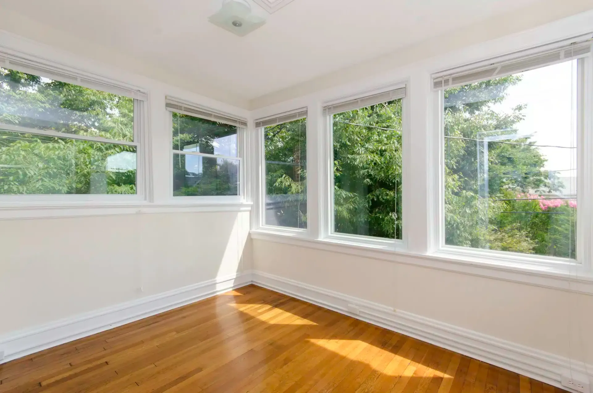 Sunroom with wood floor and several windows overlooking greenery.