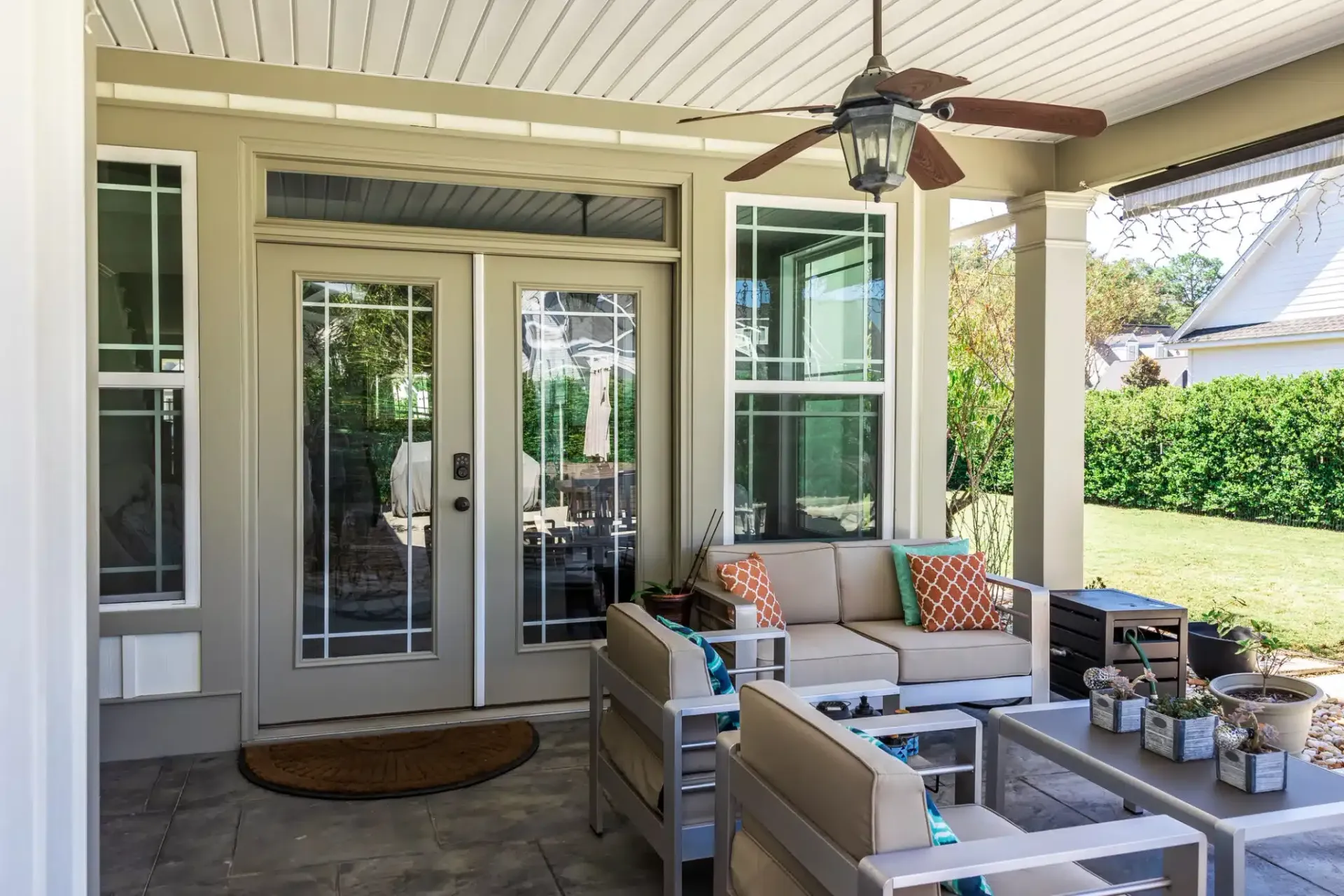 Covered outdoor patio with seating and door. Beige walls and ceiling with a ceiling fan.