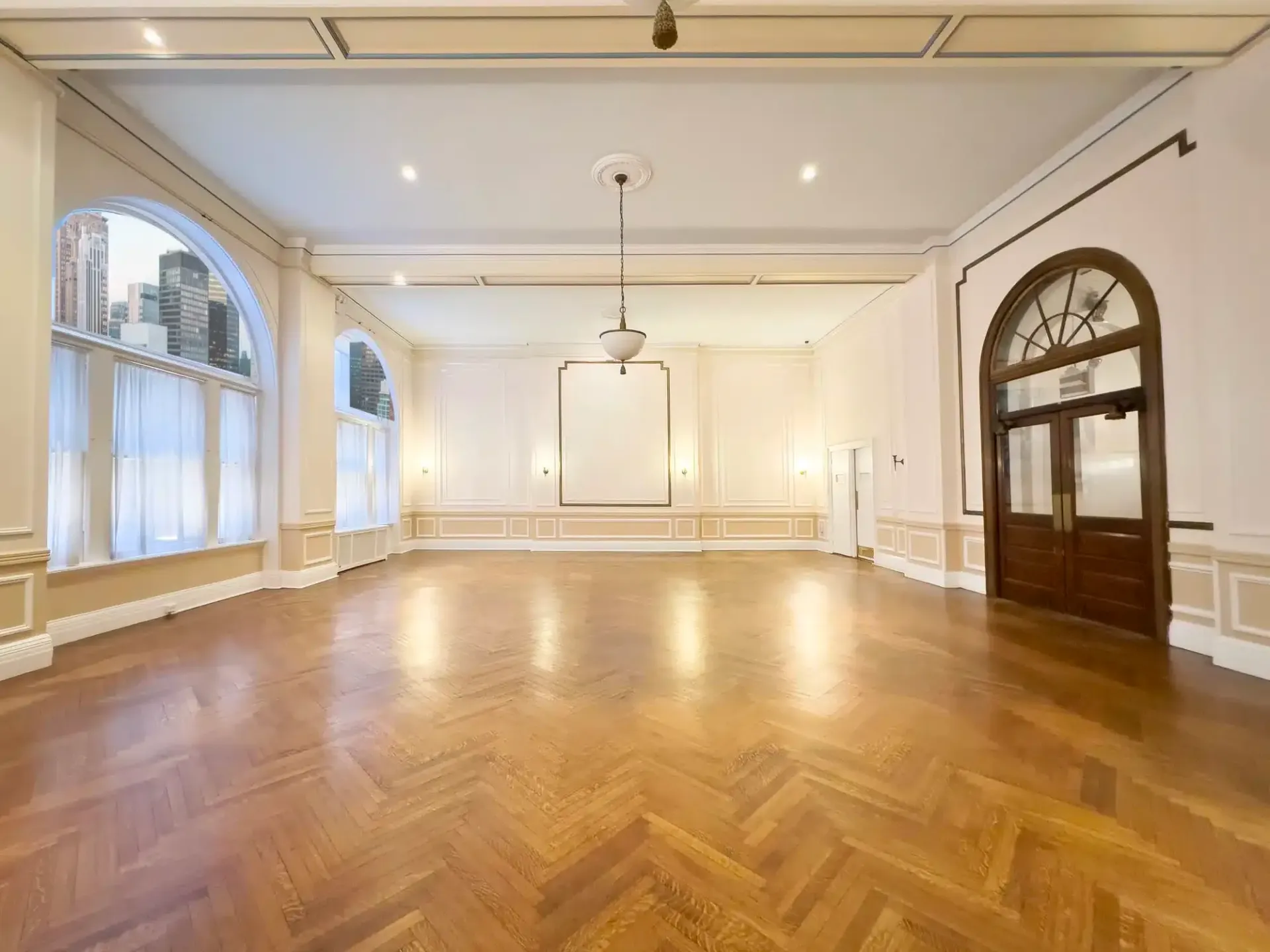 Empty ballroom with wood floor, large arched windows, and a hanging light.