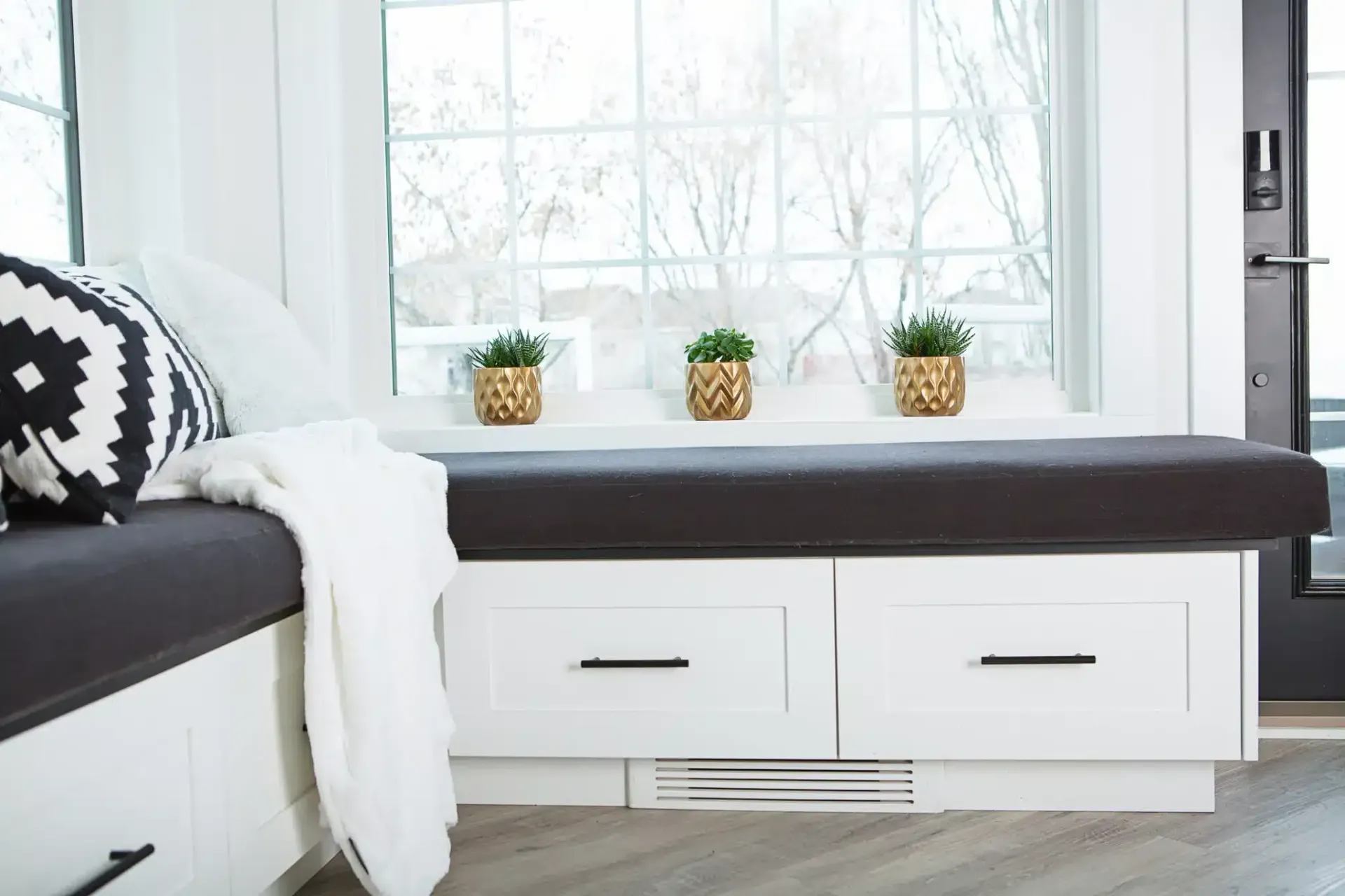 Window seat with storage drawers, dark cushion, and decorative pillows. White trim and three small potted plants.