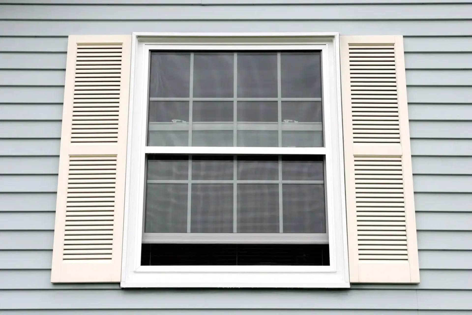 White-framed window with screen and cream-colored shutters on a blue-sided house.