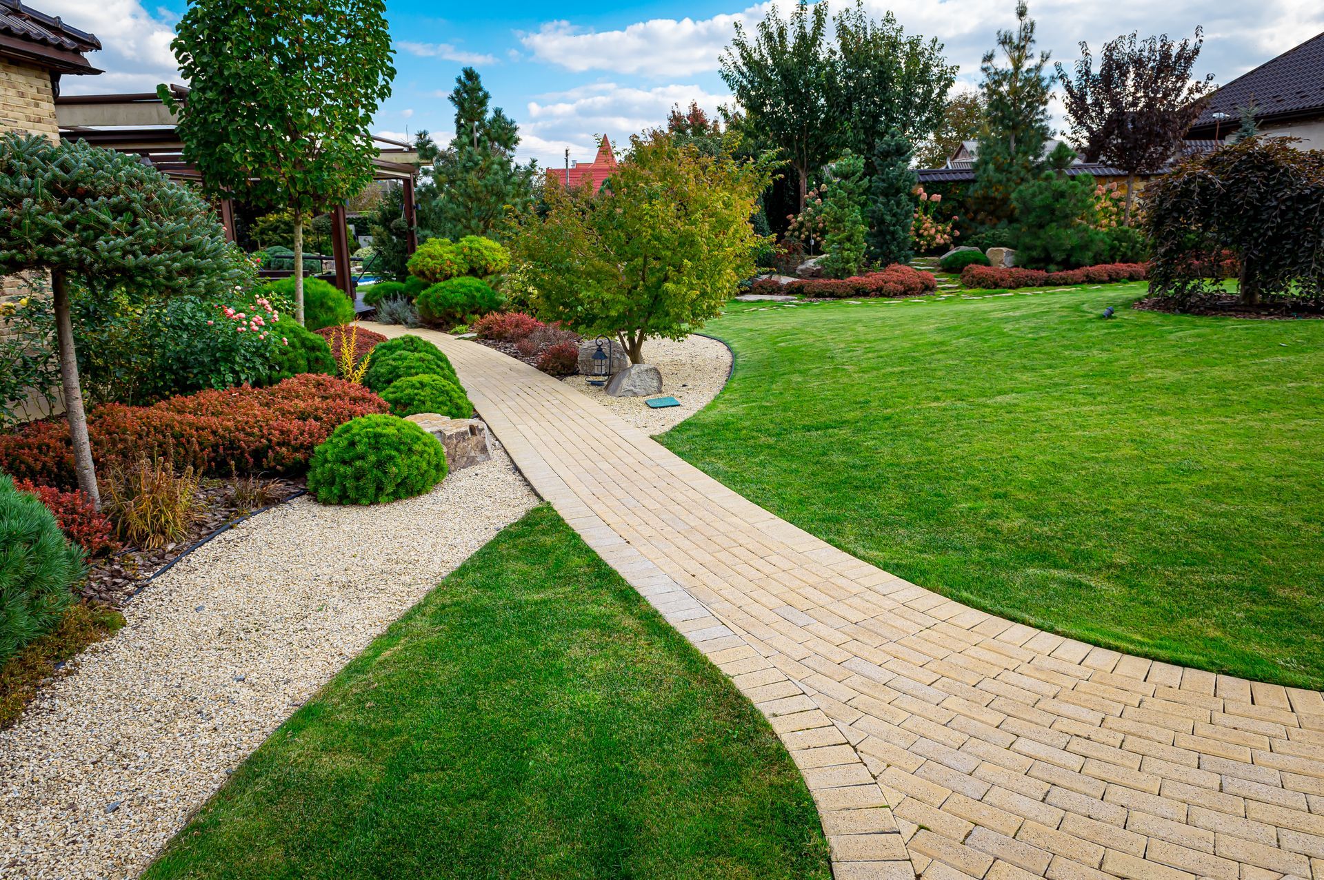 Brick pathway winding through a lush, landscaped garden with trees, shrubs, and green grass.