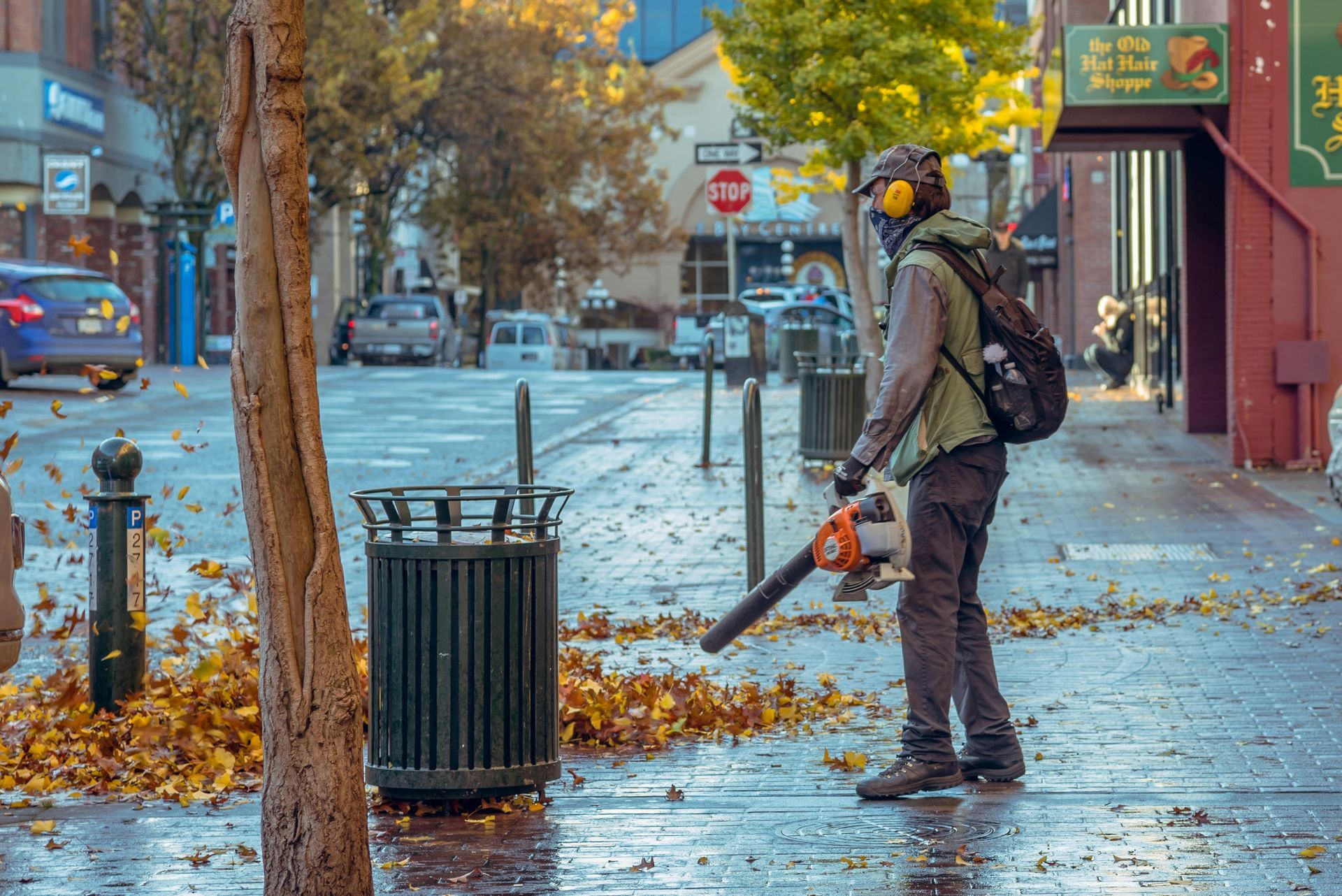 Man using a leaf blower on a wet city sidewalk, clearing fallen leaves near a trash can and trees.