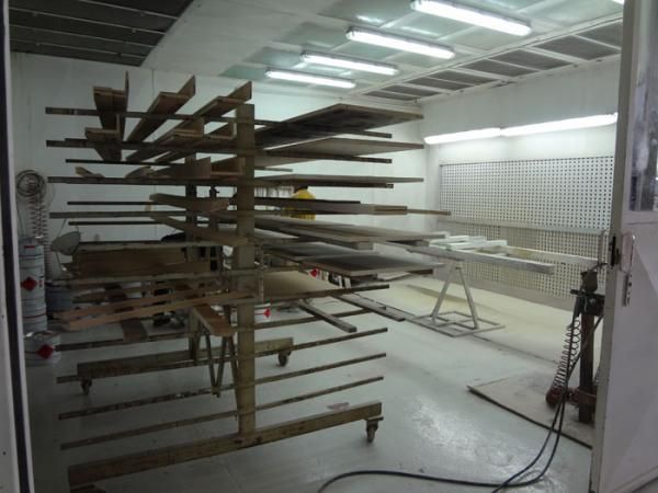 Interno di una cabina di verniciatura con ripiani in legno su cui sono appoggiate le assi, illuminati da luci soffuse.