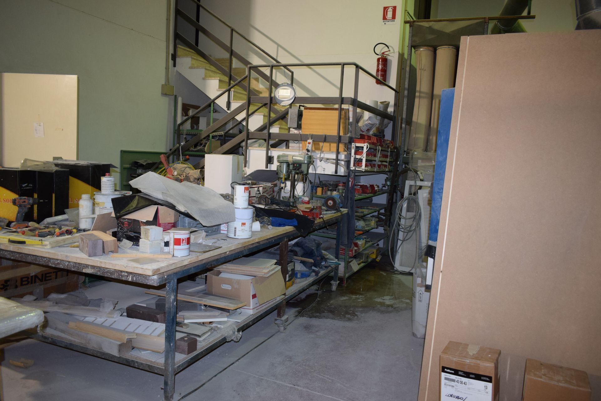 Interno dell'officina con banco da lavoro, scaffali, scale, attrezzi e materiali.